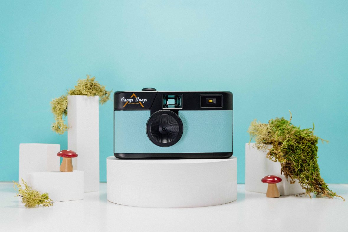 A small camera the size of a disposable camera, with a blue and black front, on a white pedestal against a blue background. There are tiny fake toadstool mushrooms as decoration.