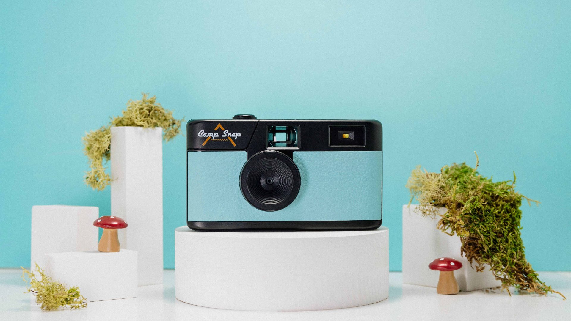 A small camera the size of a disposable camera, with a blue and black front, on a white pedestal against a blue background. There are tiny fake toadstool mushrooms as decoration.