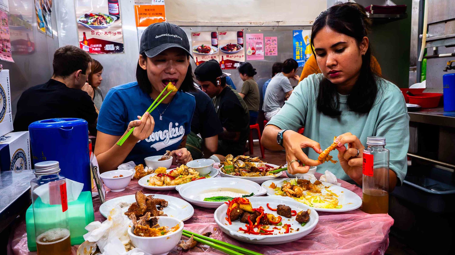 Two women eat with chopsticks at a crowded restaurant. Dishes on the table are half eaten.