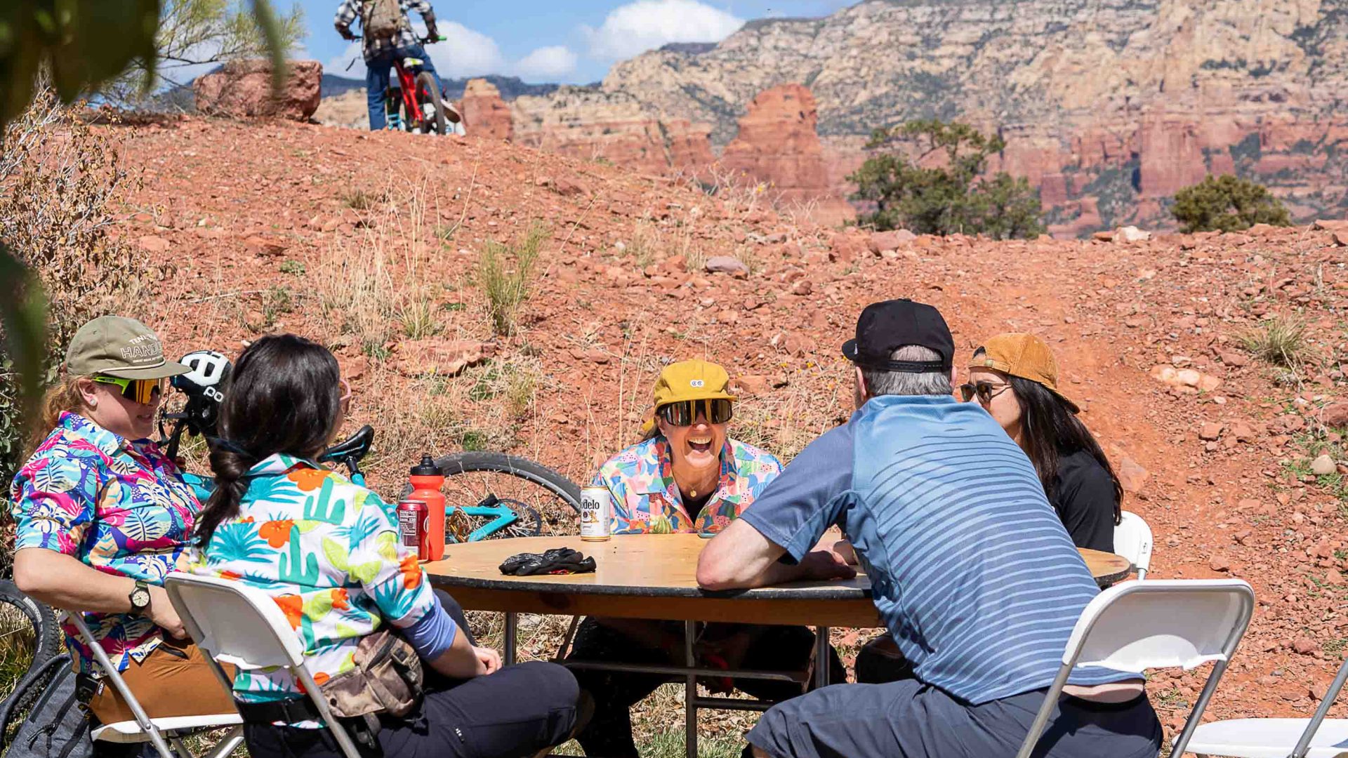 People sit around a table laughing while a mountain biker can be seen on a hill in the background.