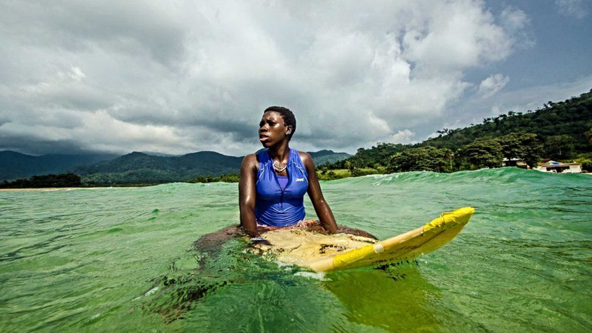 Kadiatu Kamara on her surfboard in the water. She looks away from the camera.