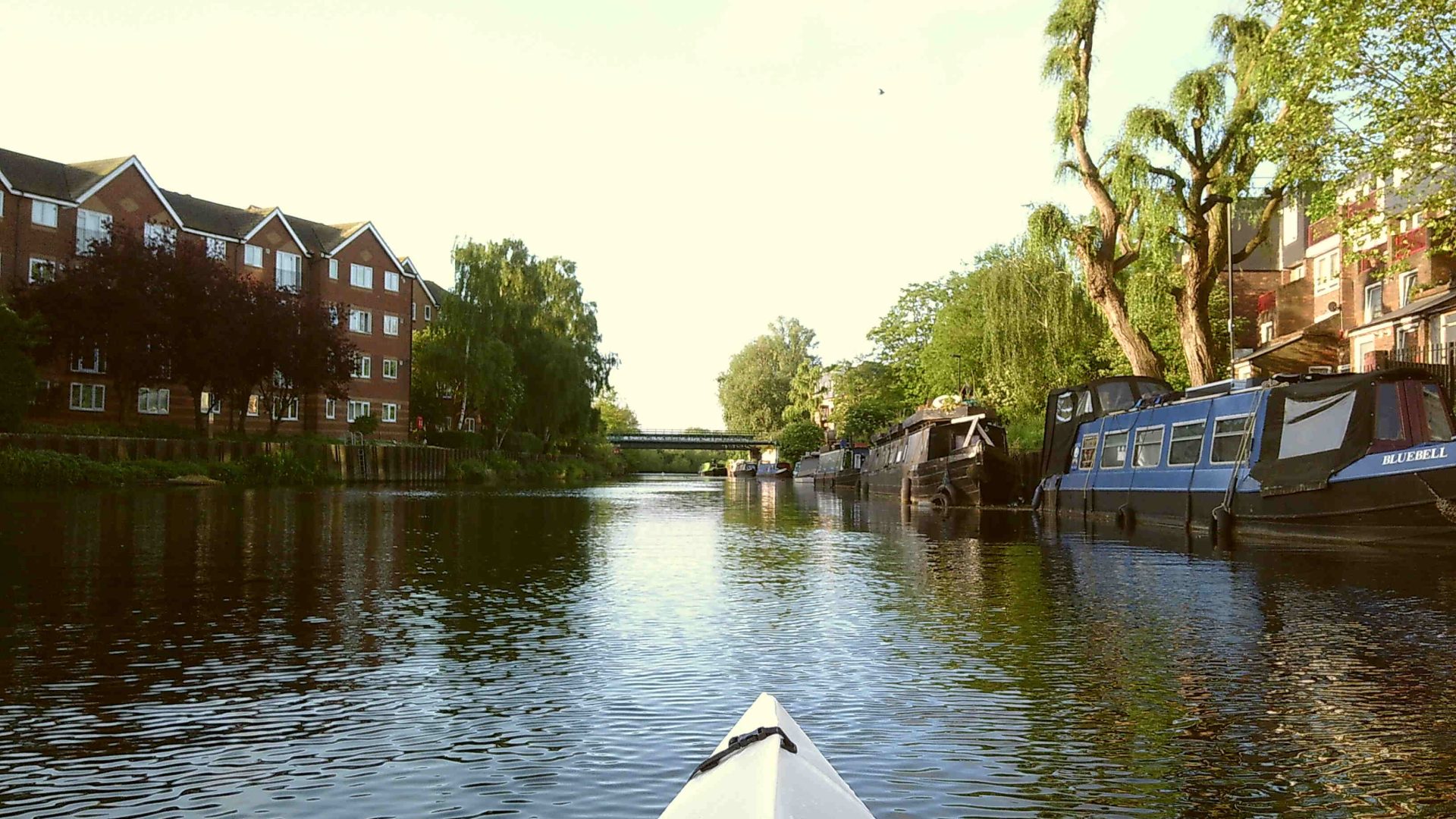 The nose of a white Oru kayak in a canal in East London, with buildings to the left and houseboats to the right.