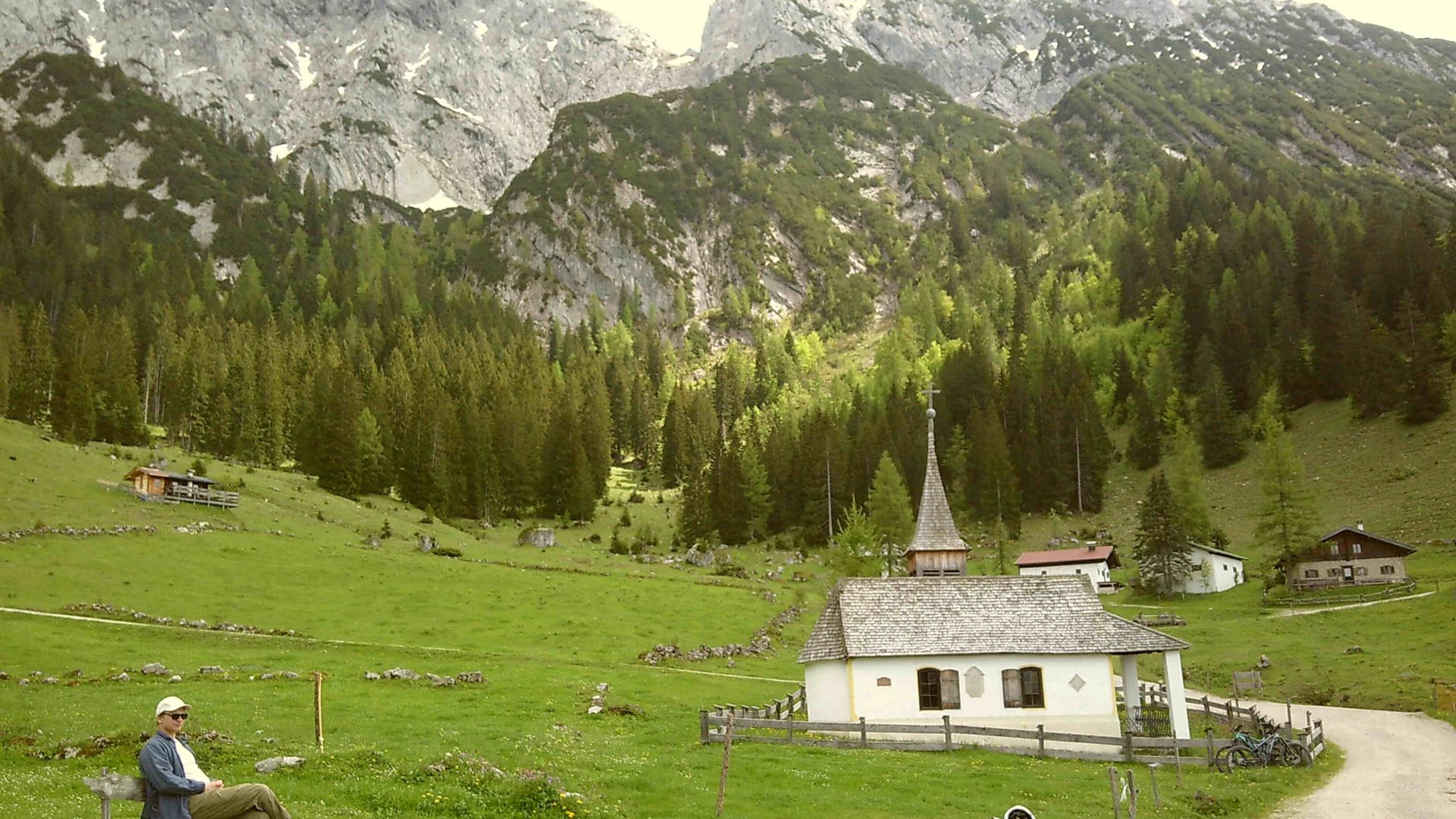 A small white church surrounded by grass, in front of a dramatic mountain view.