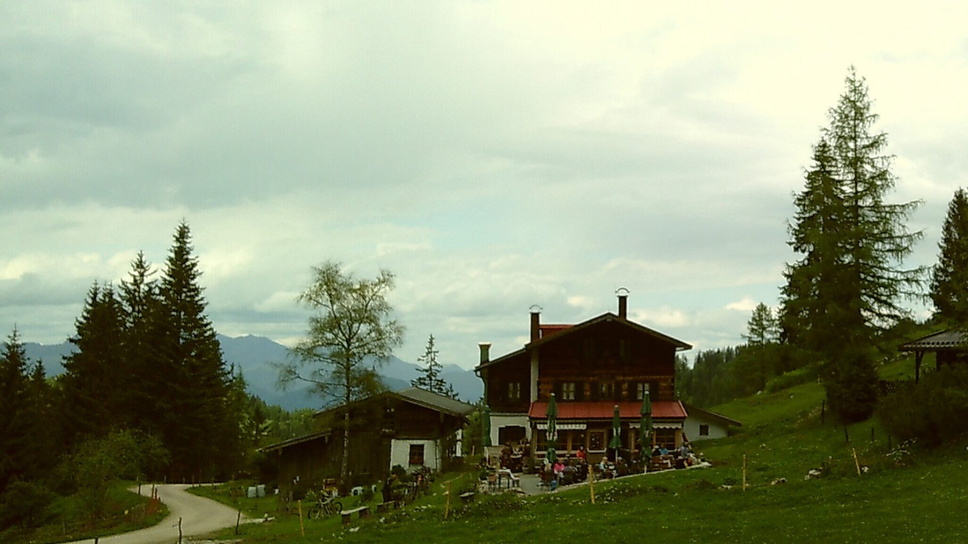 A mountain hut surrounded by pine trees with a mountain view in the distance. A path can be seen to the left.