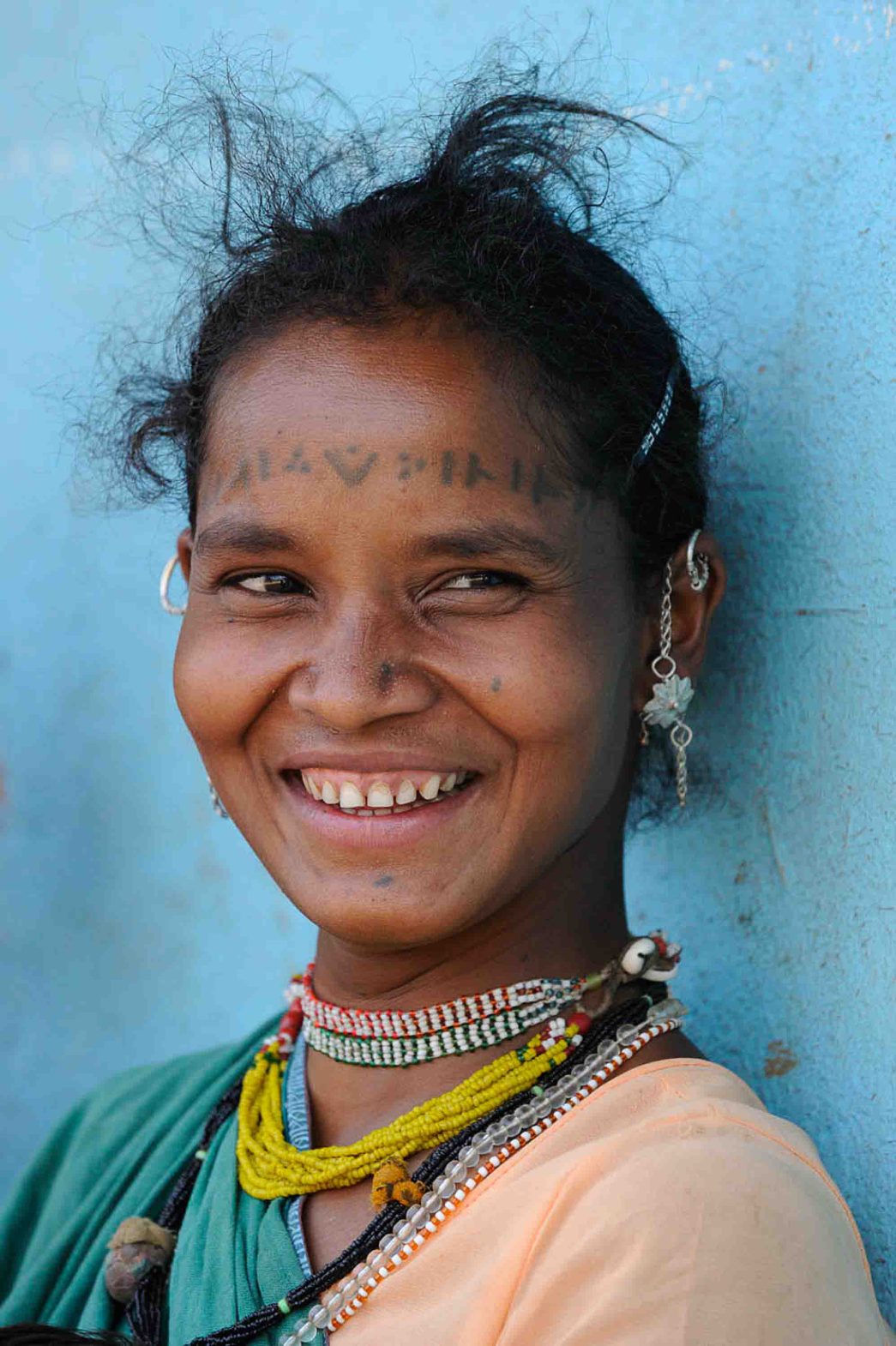 A woman with tattoos on her forehead and a necklace smiles.