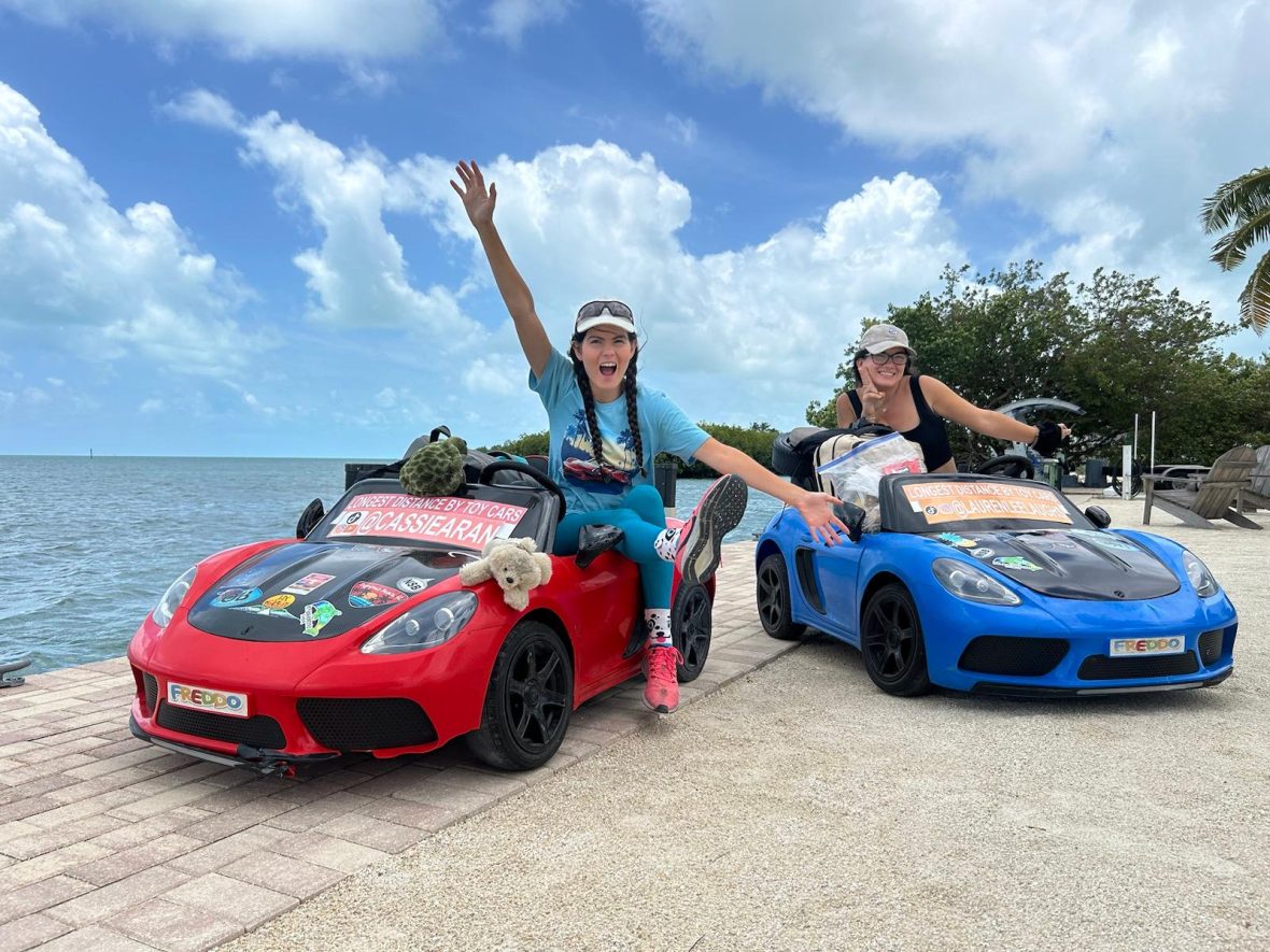 Two women pose with toy cars on a beach in Florida