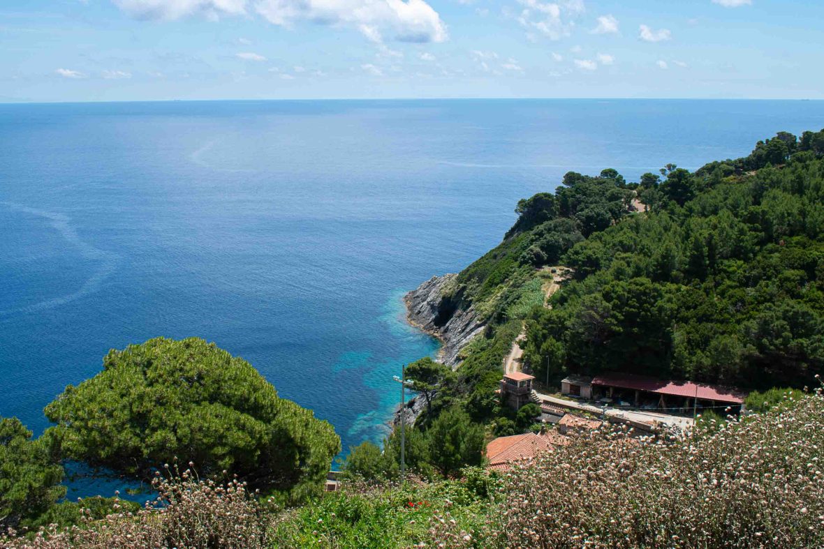 A view out over blue sea, green hill and a few buildings.