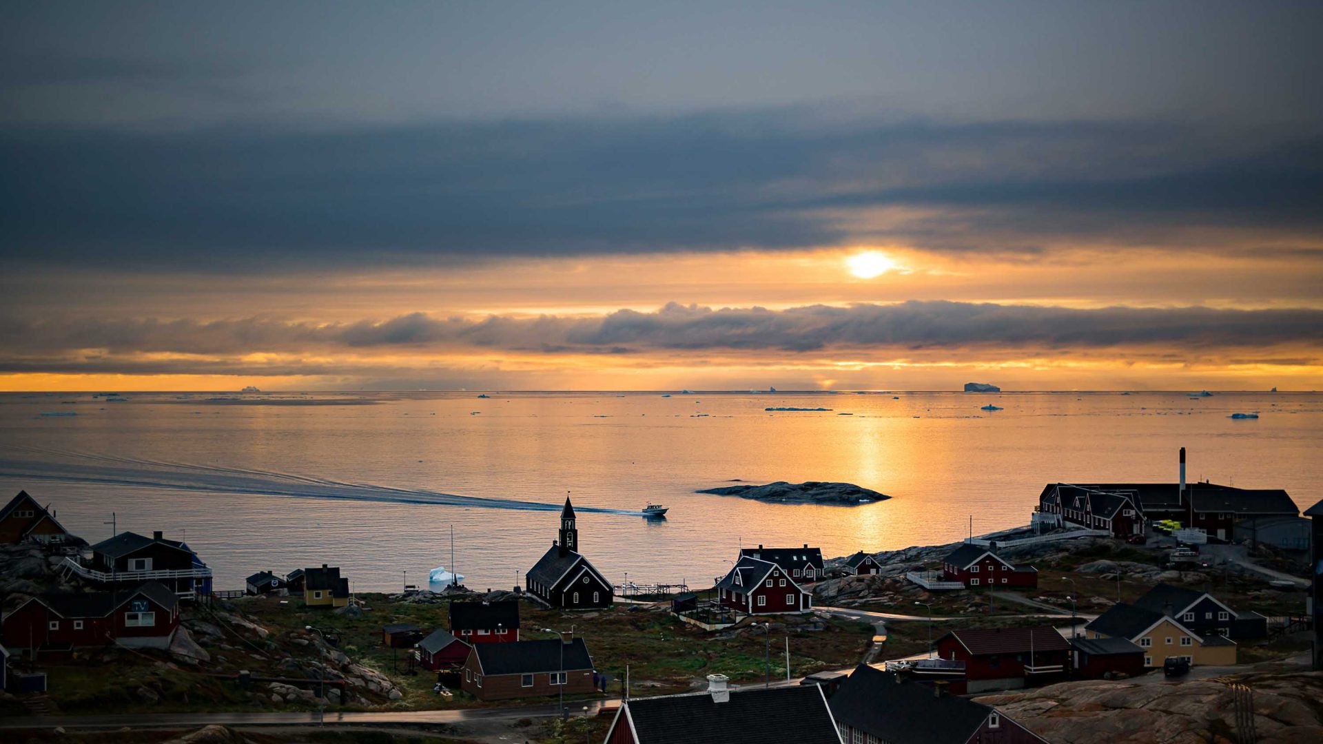 A low sun in the sky at midnight. It casts light over water and houses.