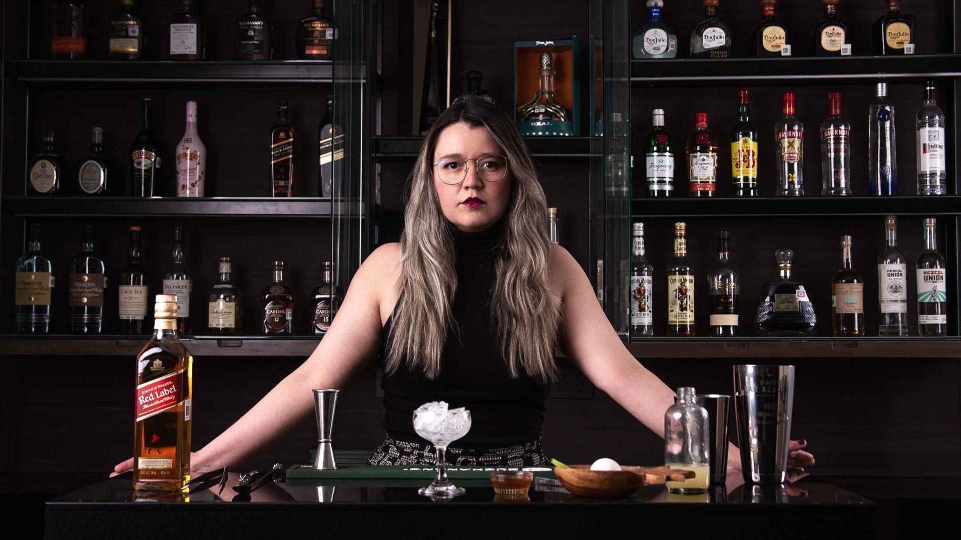 A woman stares at the camera with lots of bottles at a bar behind her.