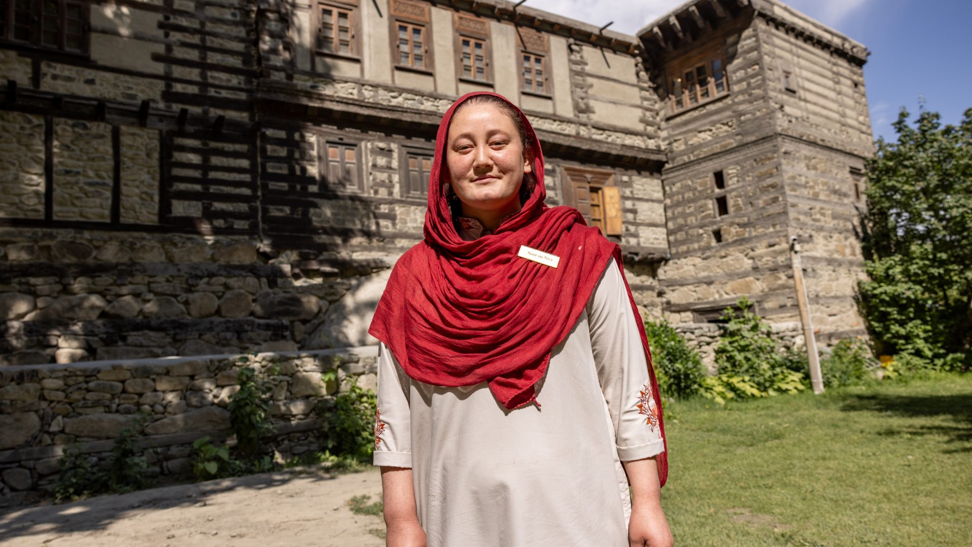 A woman in a long dress and scart stands smiling in front of a stone building