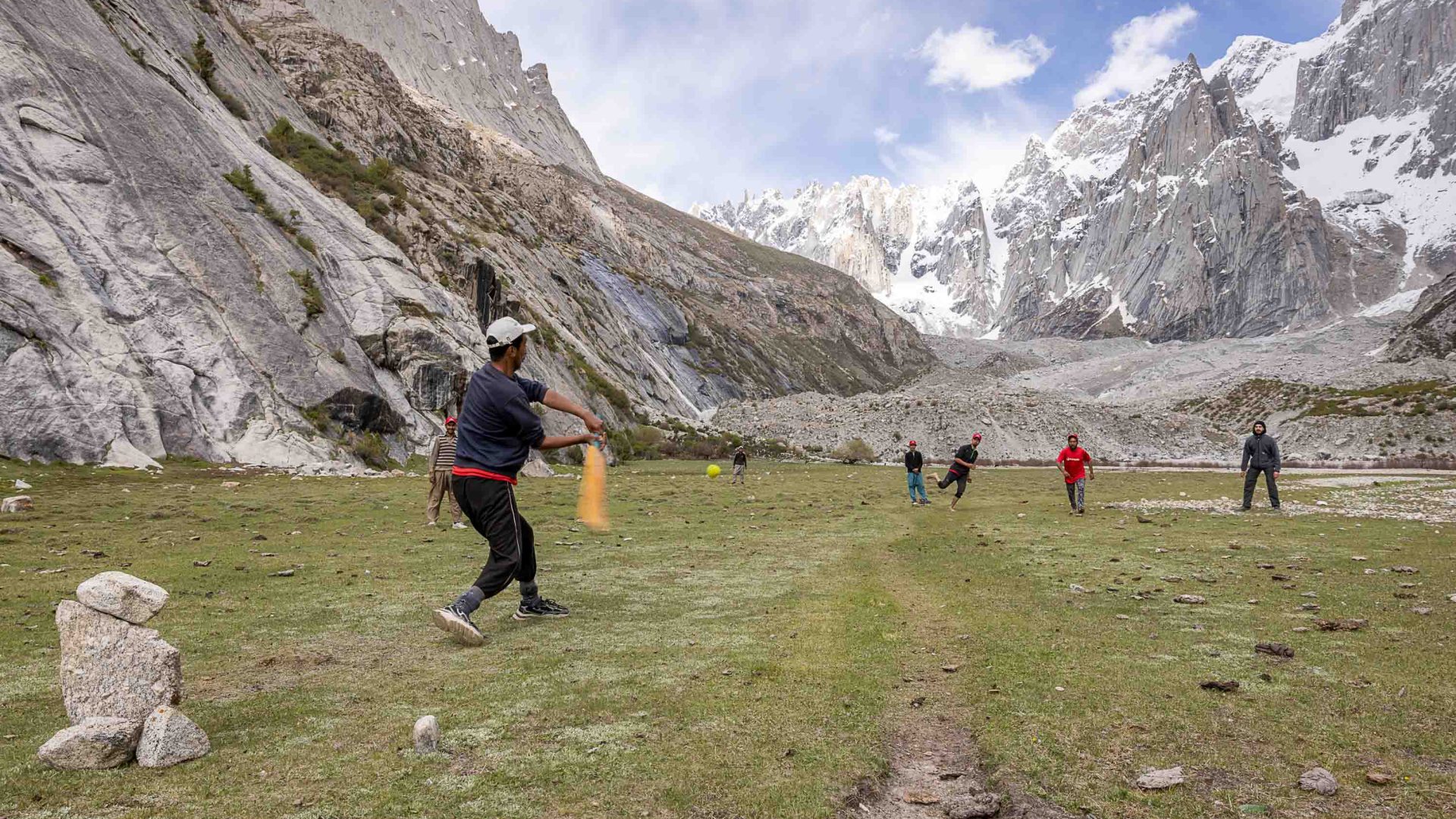 Travellers play cricket on a grassy area below mountains.