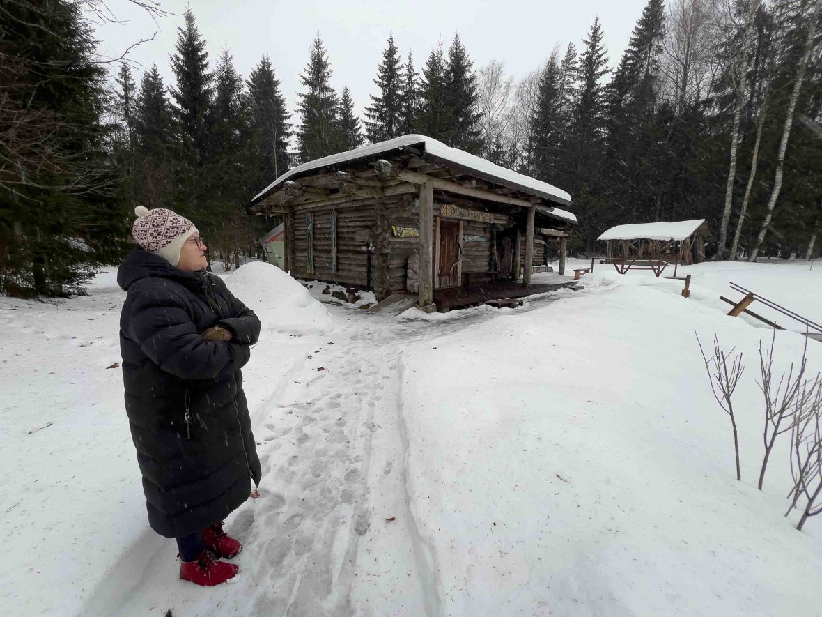 A woman stands in the snow in front of a smoke sauna.