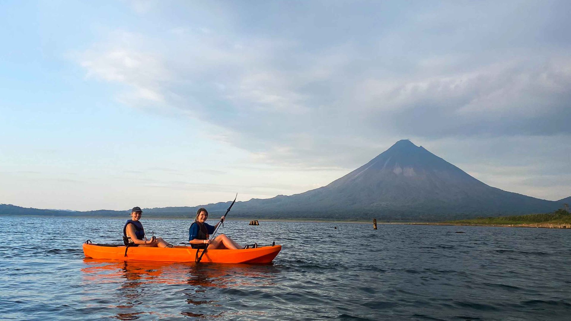 Two women are kayaking on a lake with a volcano in the background.