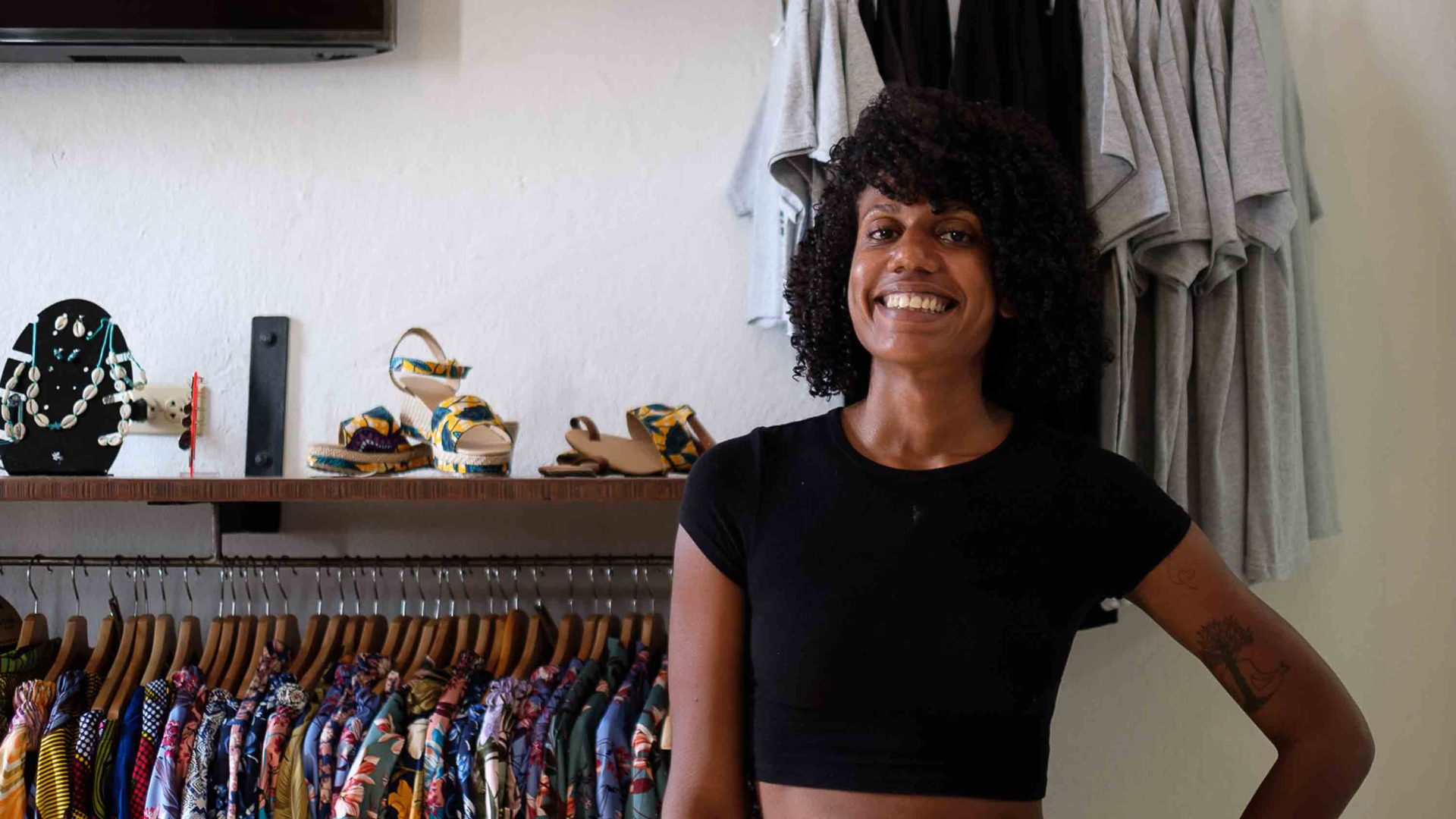 A portrait of a woman smiling in front of a row of shirts and other things for sale.