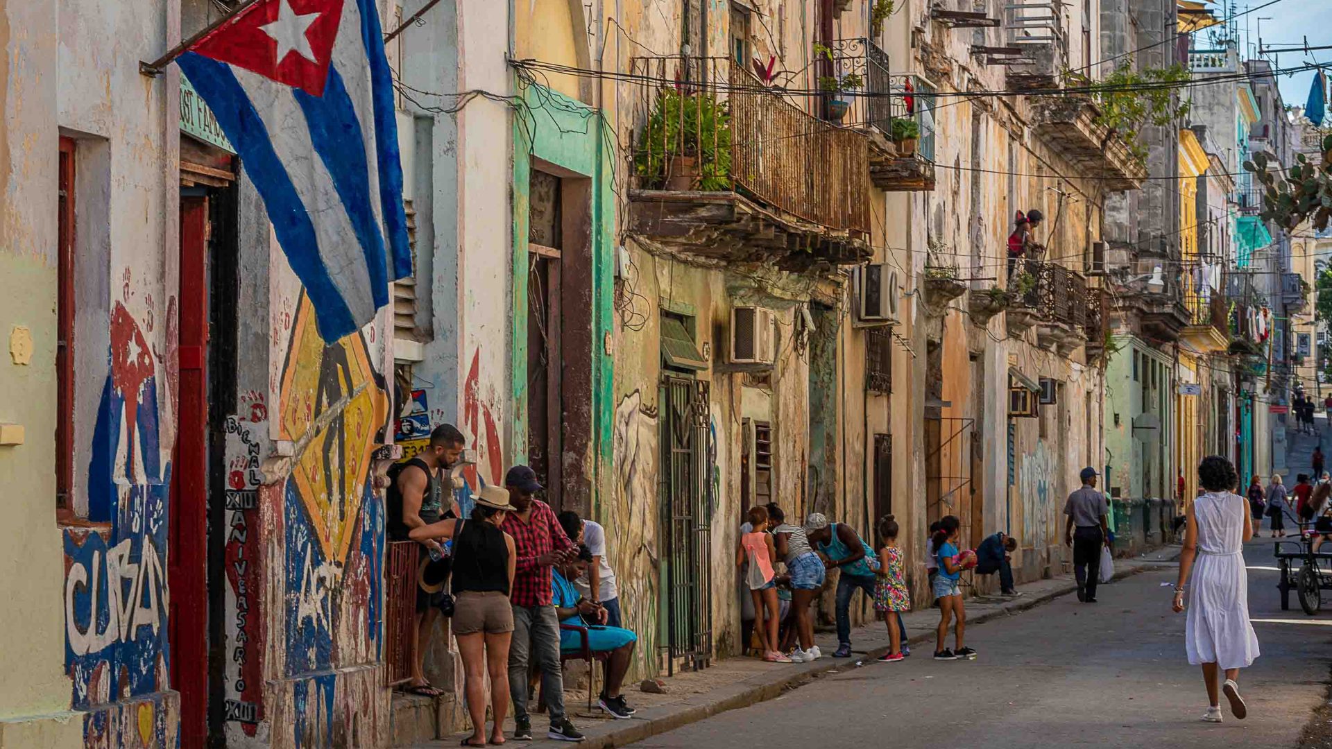 A Cuban flag hangs from one of the colourful Havana houses. People mill about in the streets.