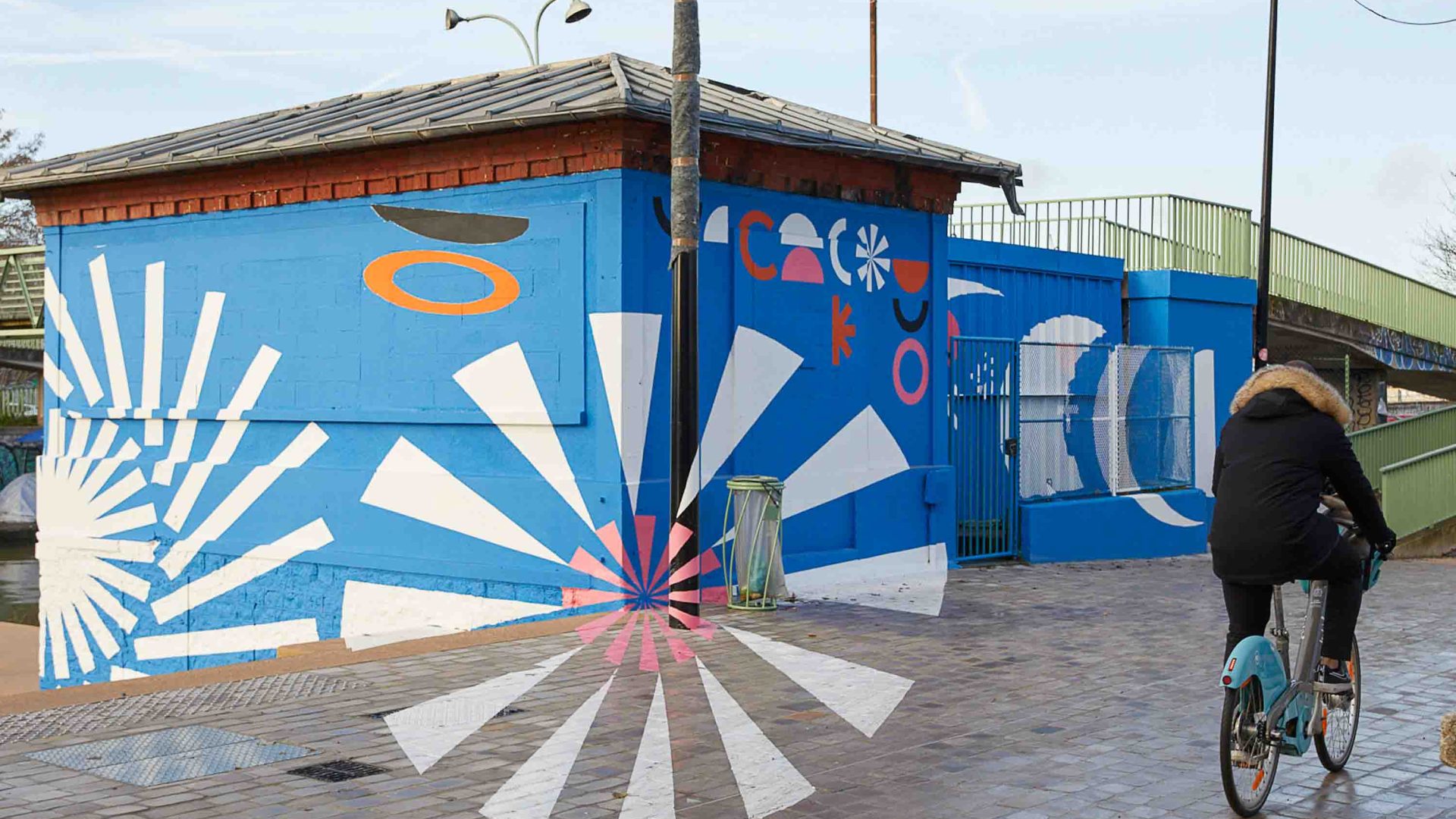 A person cycles on a cycle path, passing by a street art mural that is mostly blue.