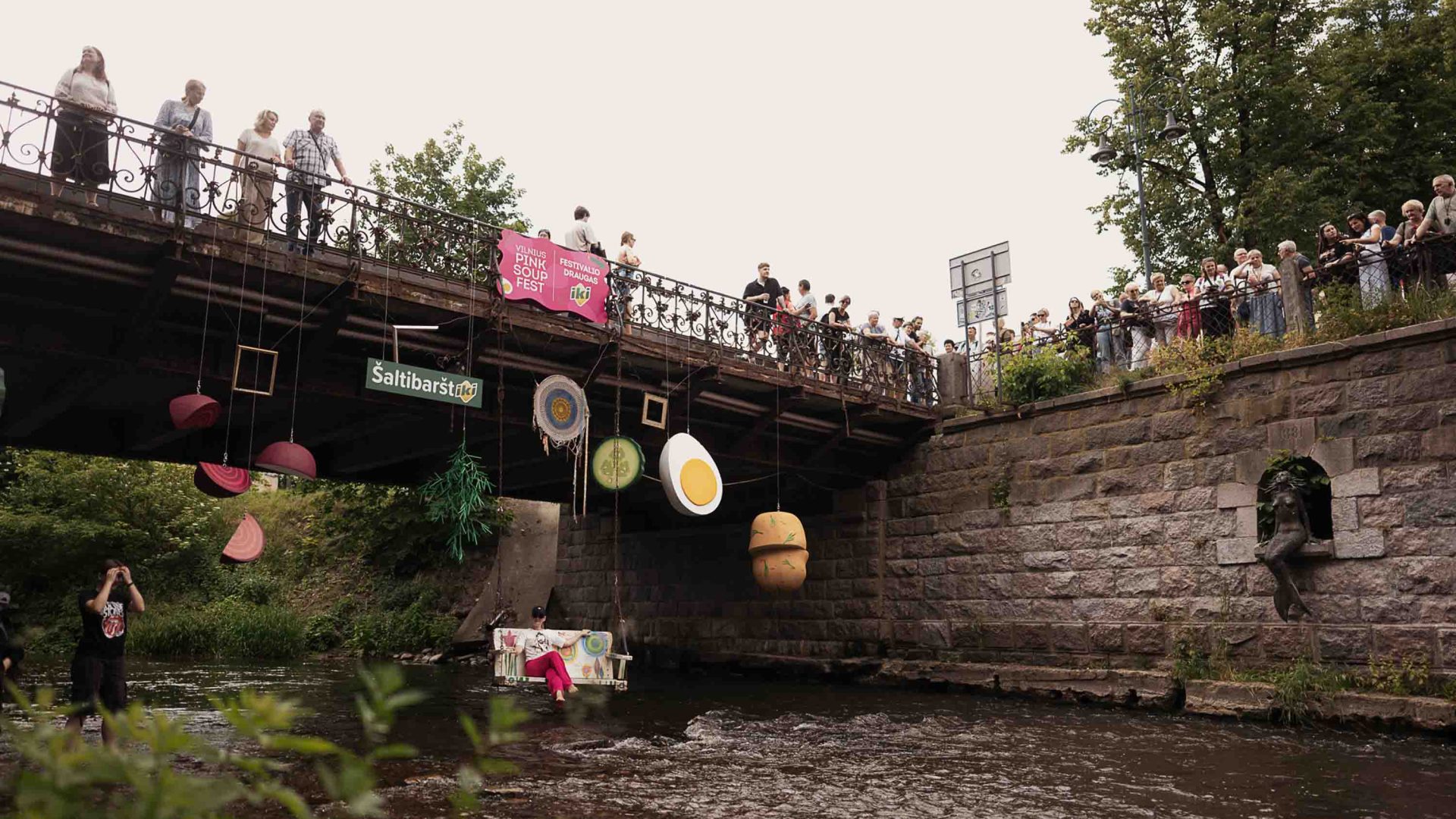 Spectators watch from a bridge as people dance in the water below. The bridge is adorned with ingredients from the soup.