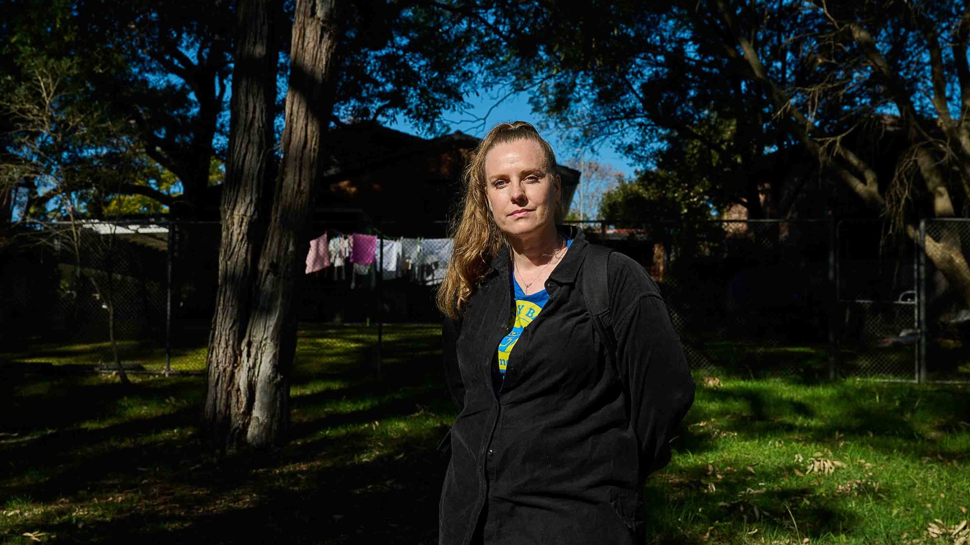 A woman stands in a tree covered back yard which has washing on the washing line.