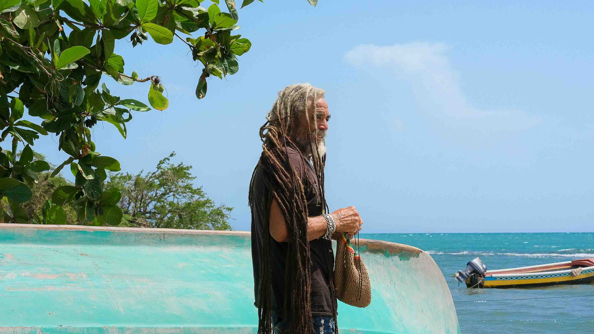 A man with dreadlocks stands by an upturned boat by the sea.