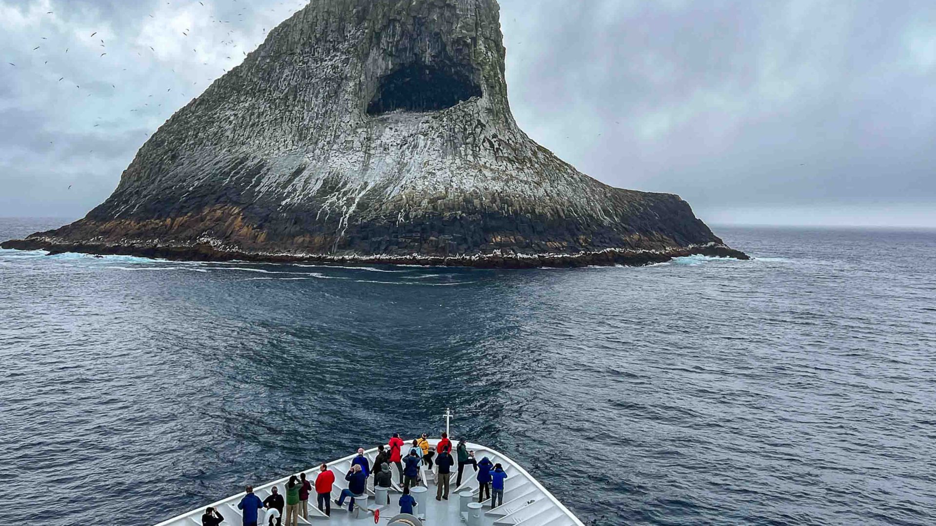 People on board a ship look at a pyramid shaped island.