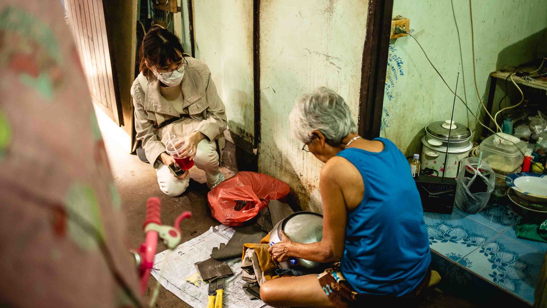 Two women sit on the floor of a home kitchen while one produces music.