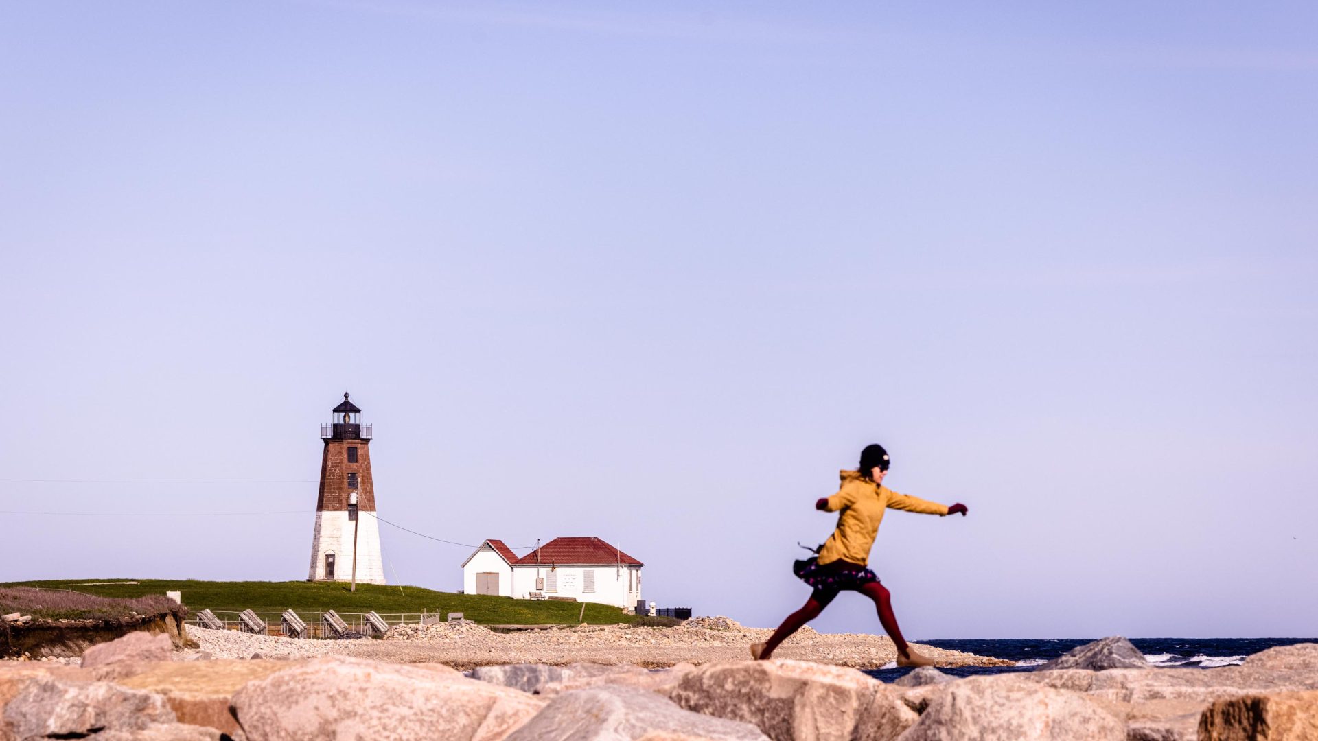 A woman hops across a stone breakwater in front of the Point Judith Lighthouse in Narragansett, Rhode Island.