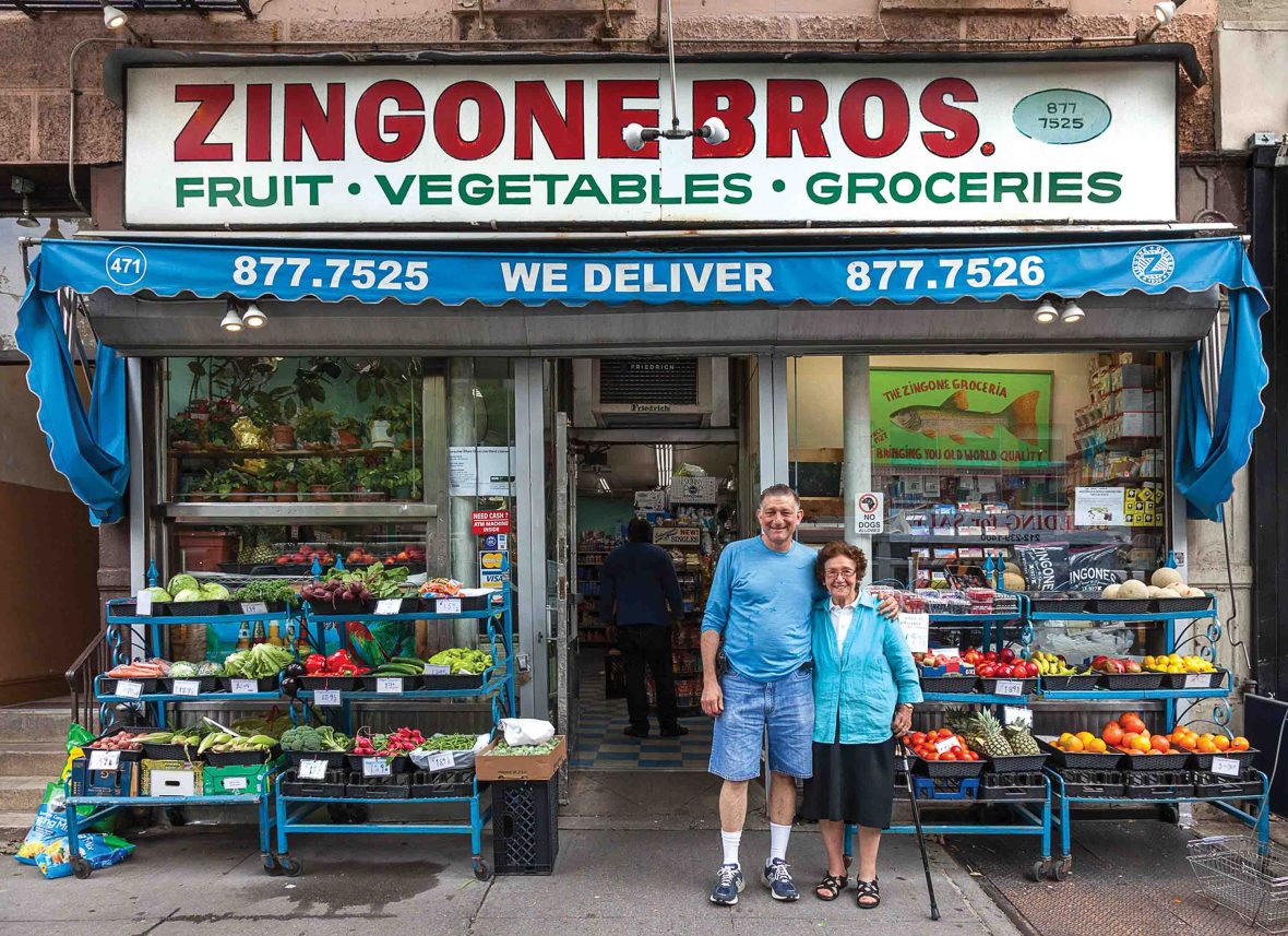 A couple stand together outside their store. It has displays of fruit and vegetables.