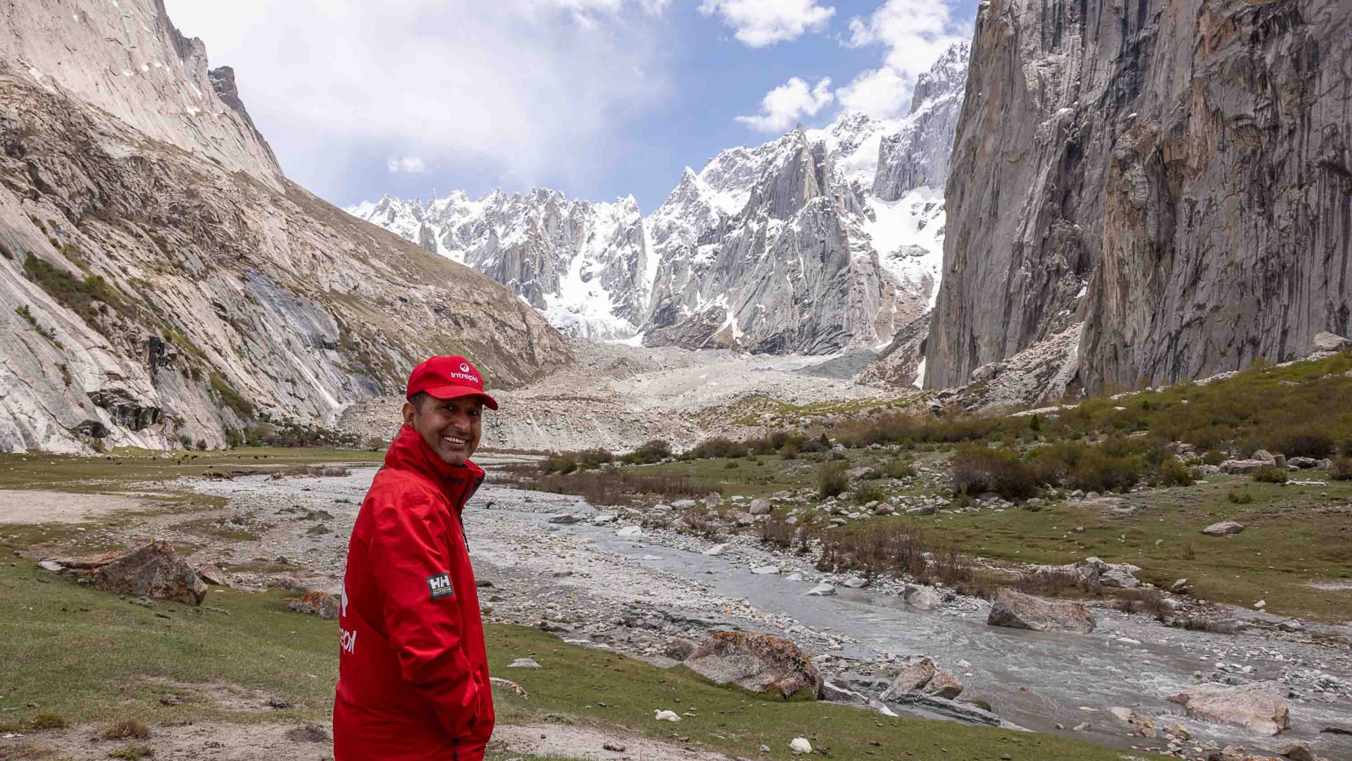 A man smiles to camera. He stands in front of mountains and a river.