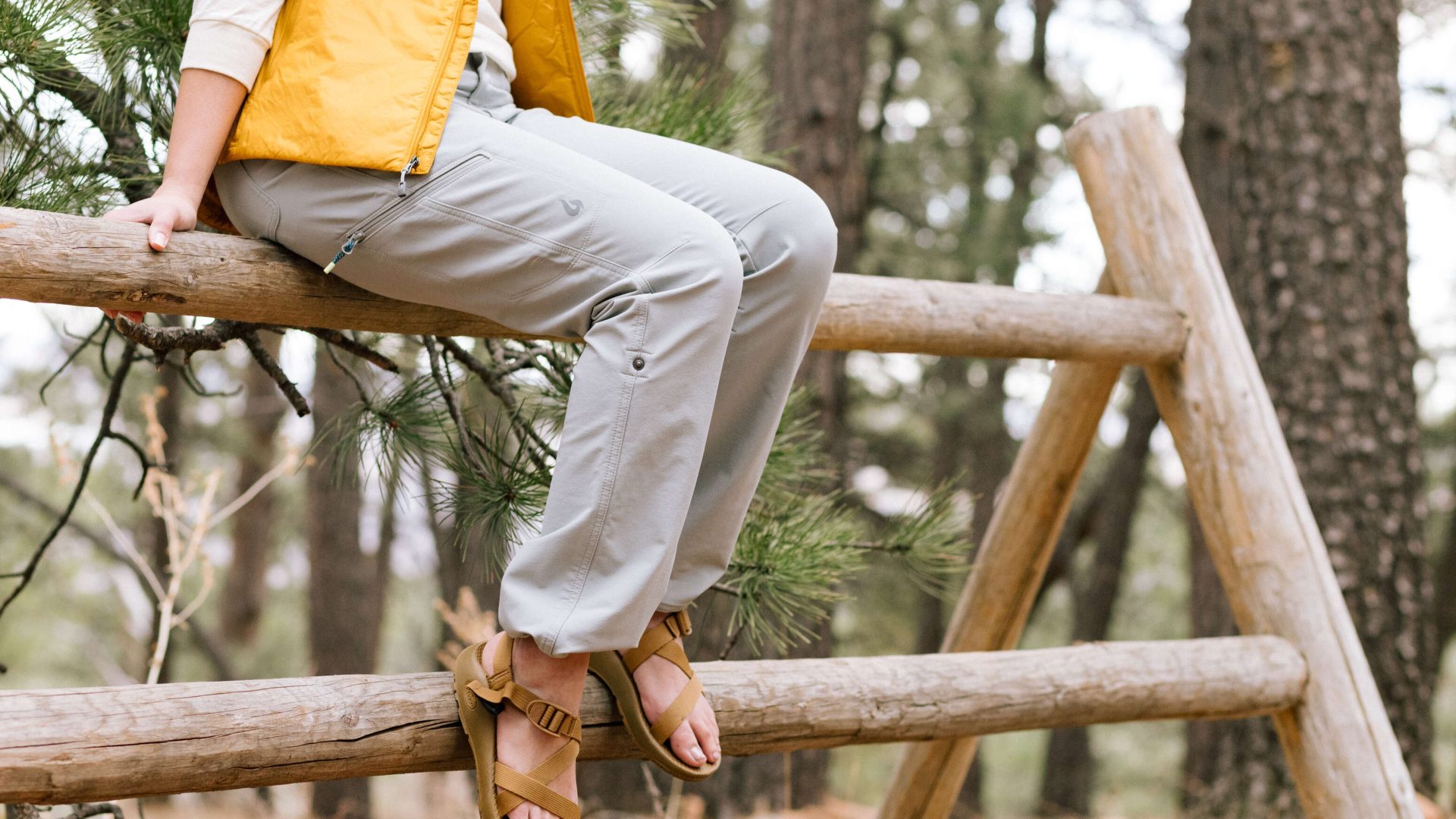 A hiker wearing a yellow vest and gray Gnara hiking trousers while sitting on a wooden fence.