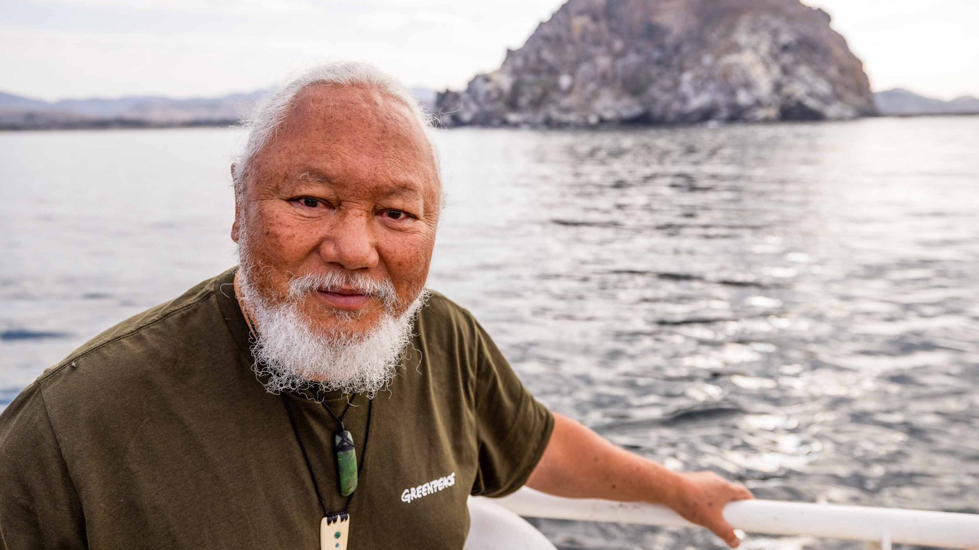 A man has a serious expression as he looks to camera. He's on a boat going in the direction of an island.