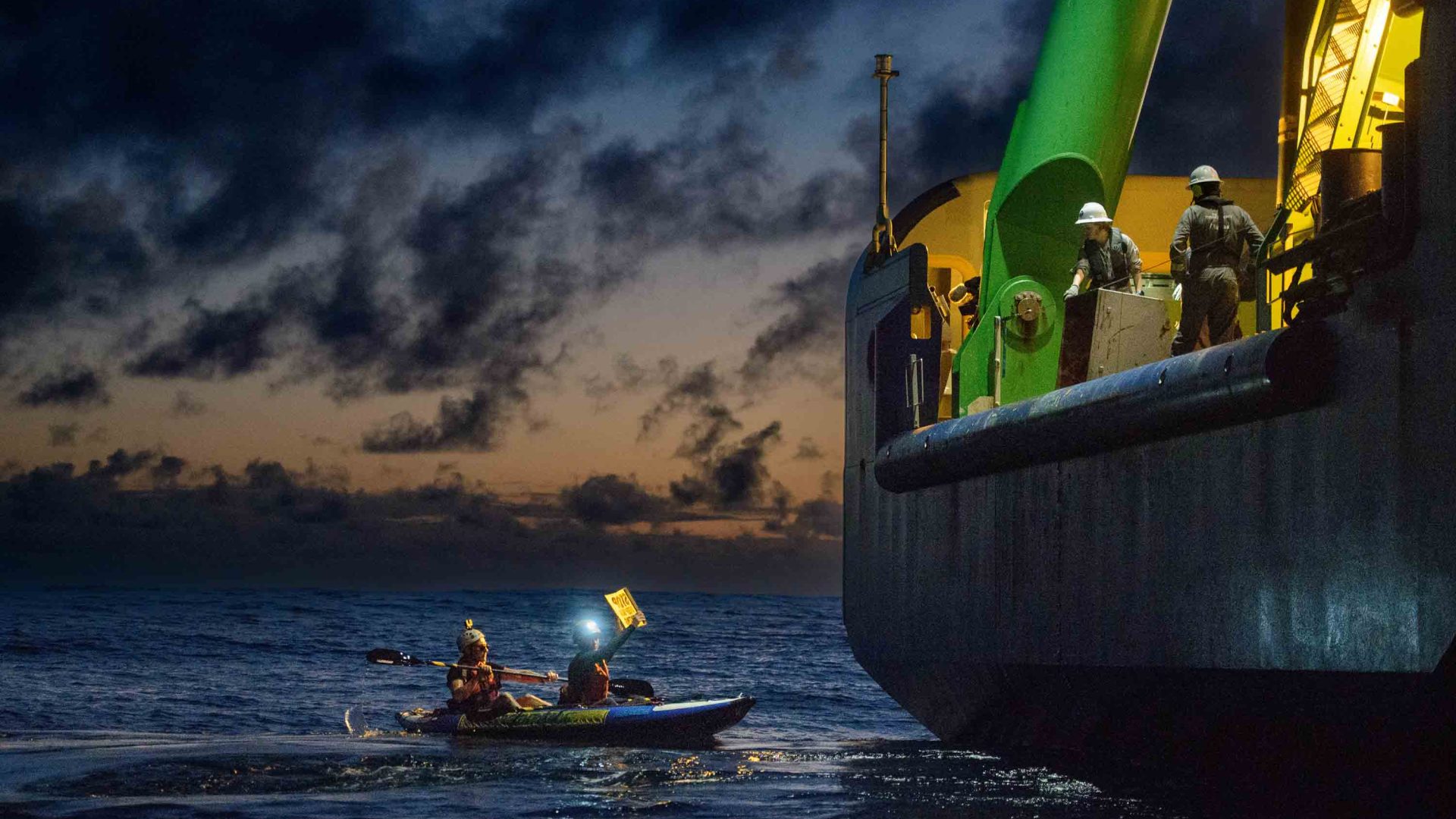 A small boat holds up a sign at night to workers on a larger ship.