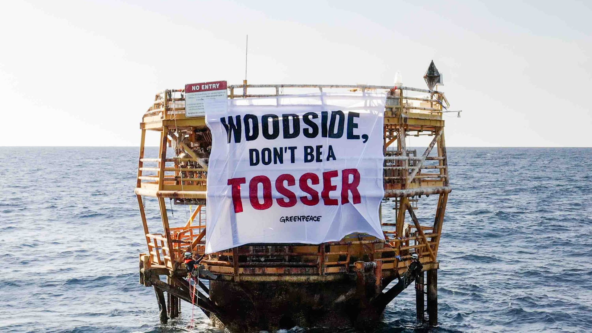 Greenpeace activists climb up an old oil tower which has a sign that reads 'Woodside don't be a tosser'.