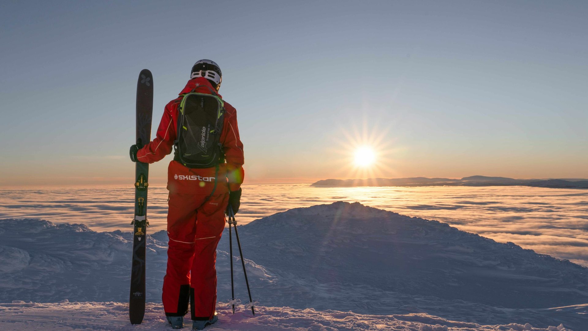 A person in red ski gear faces away from the camera, toward a sunrise as seen from the top of a mountain.