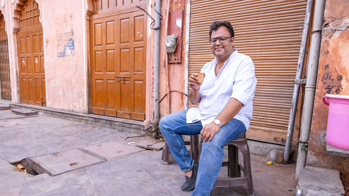 A man sits on a stool on a street drinking chai