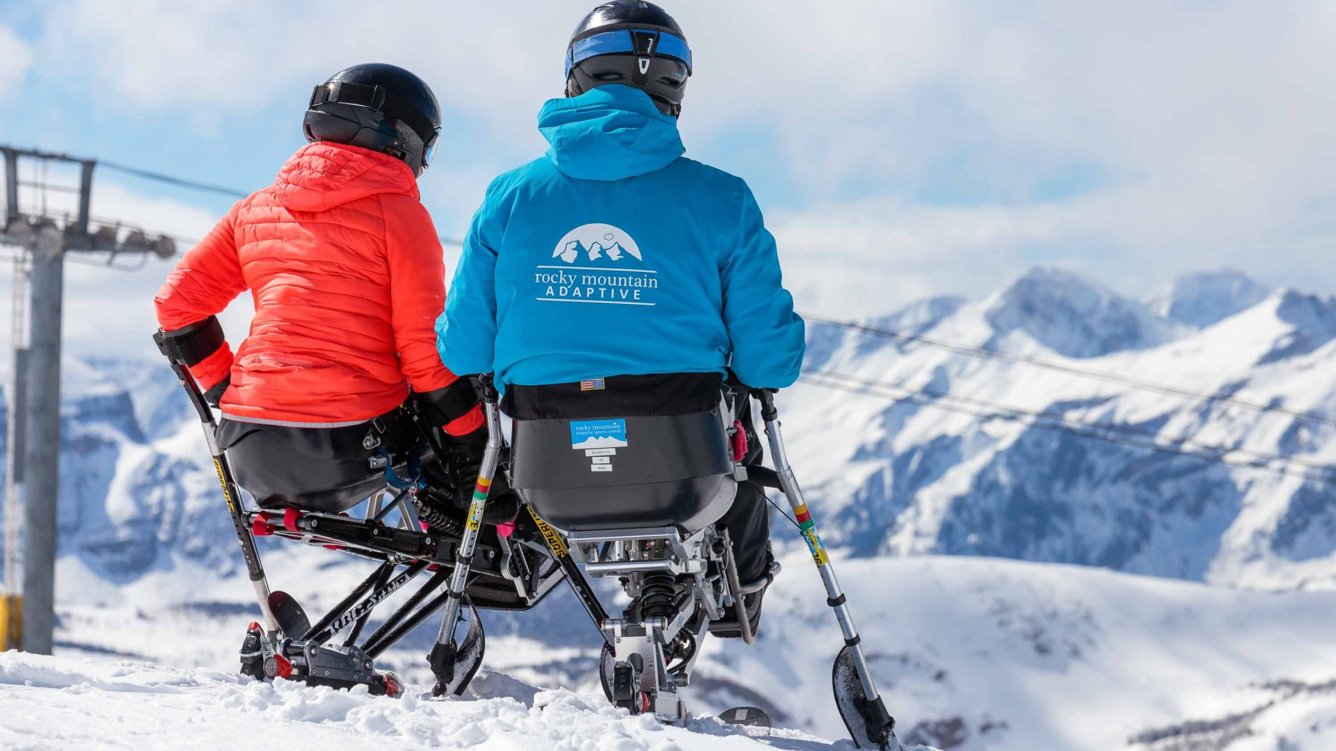 Two people in sit skis look out over snowy mountains.