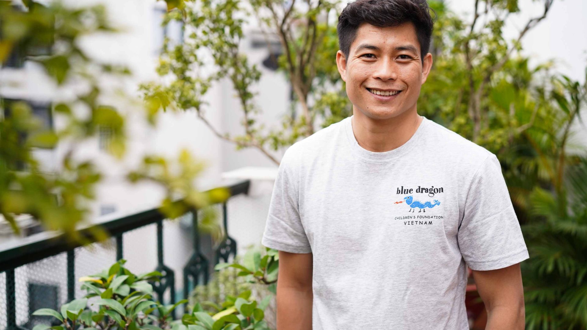 A man in a white t shirt smiles against a leafy background.