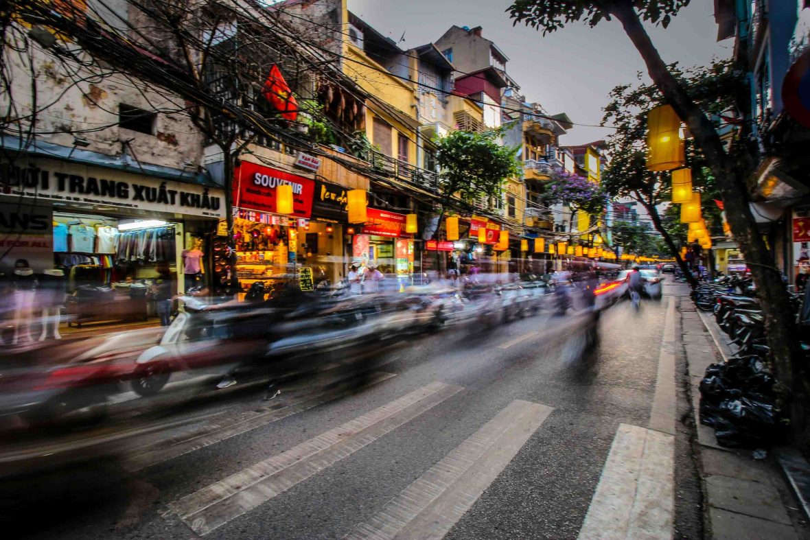 Cars are blurred as they drive past lit up shop fronts.