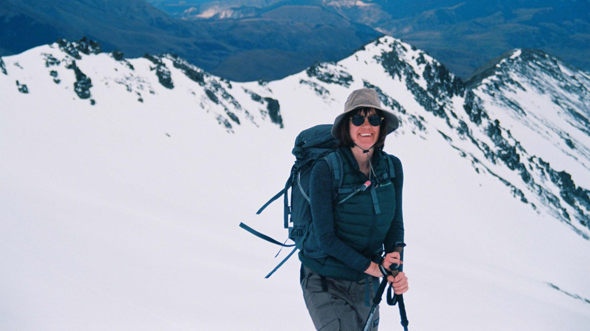 A woman in a hat and sunglasses stands in front of a snowy mountain.