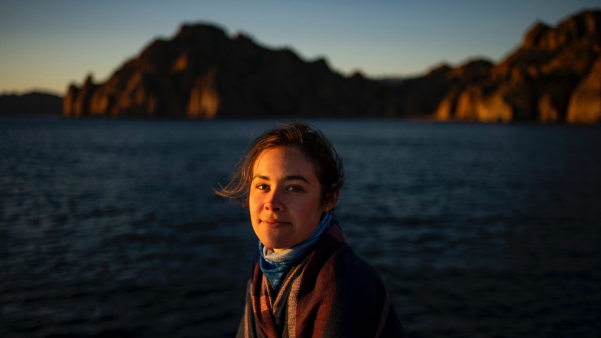 A woman with soft light on her face, water and mountains behind her.