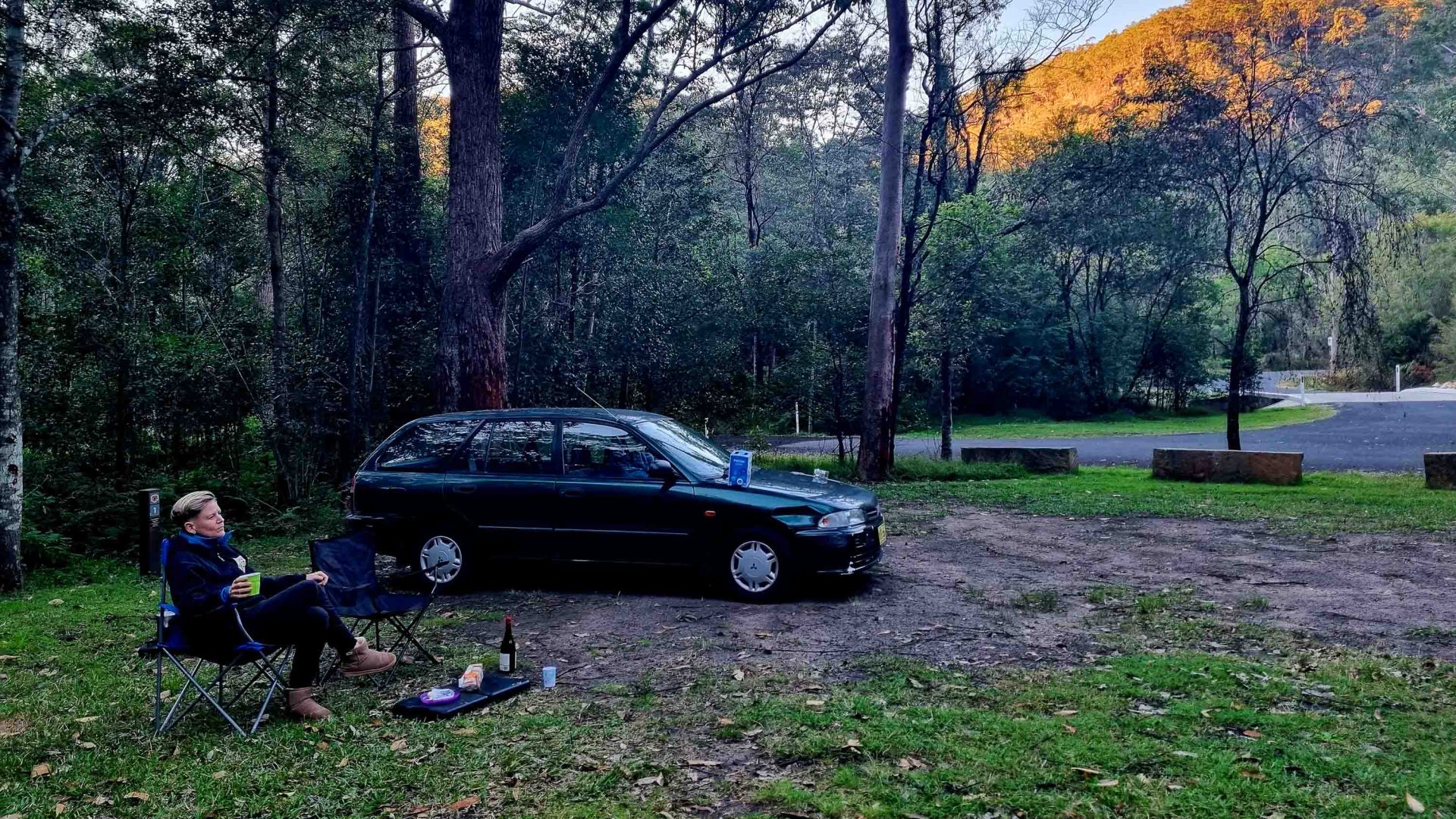 A person sits in a chair with a drink, alongside a car as the sun gets soft on the bush beside them.