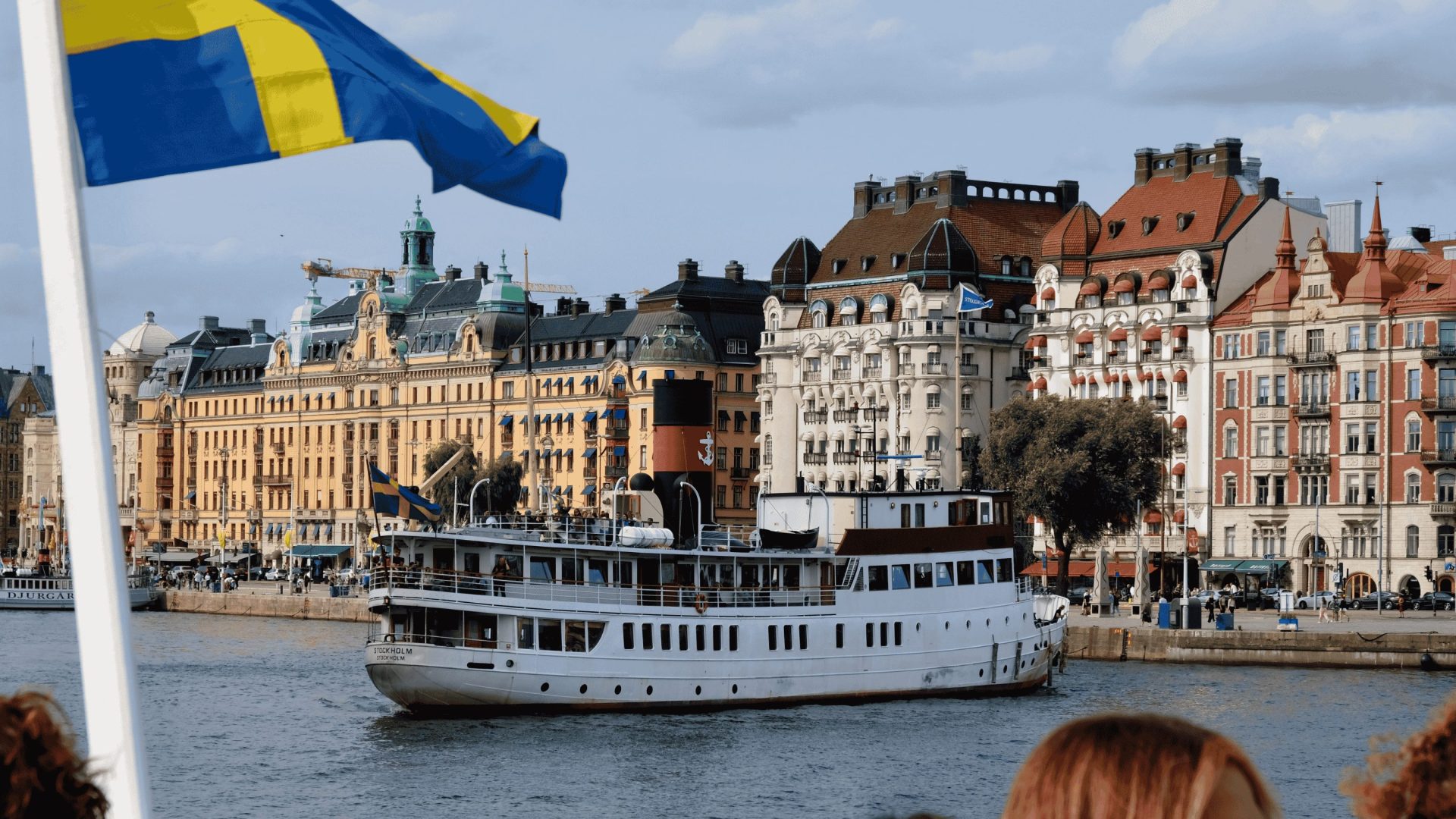 A large ferry leaves port in Stockholm, with historic buildings in the background. A Swedish flag is in the foreground.