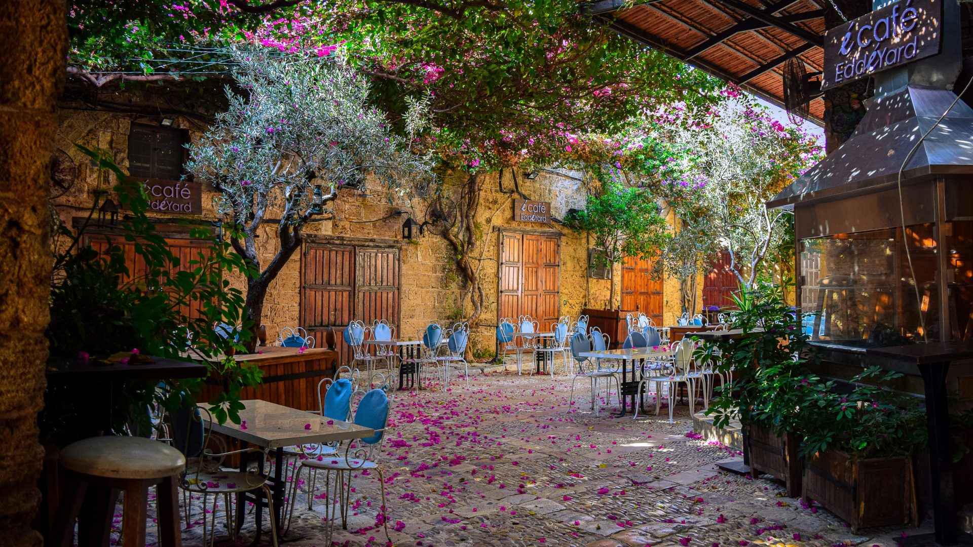 Tables and chairs in a souk that has purple flowers on the ground.