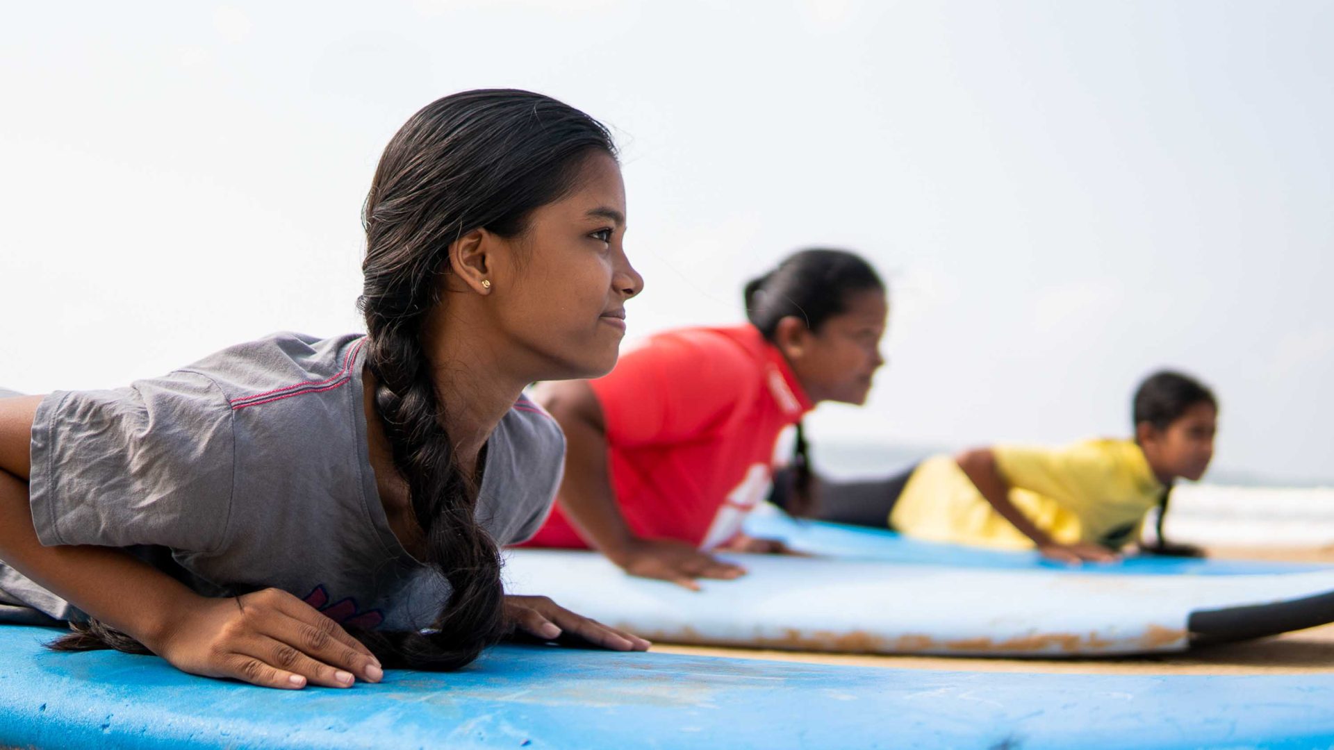 A row of women lie on boards on the sand as they learn to surf.