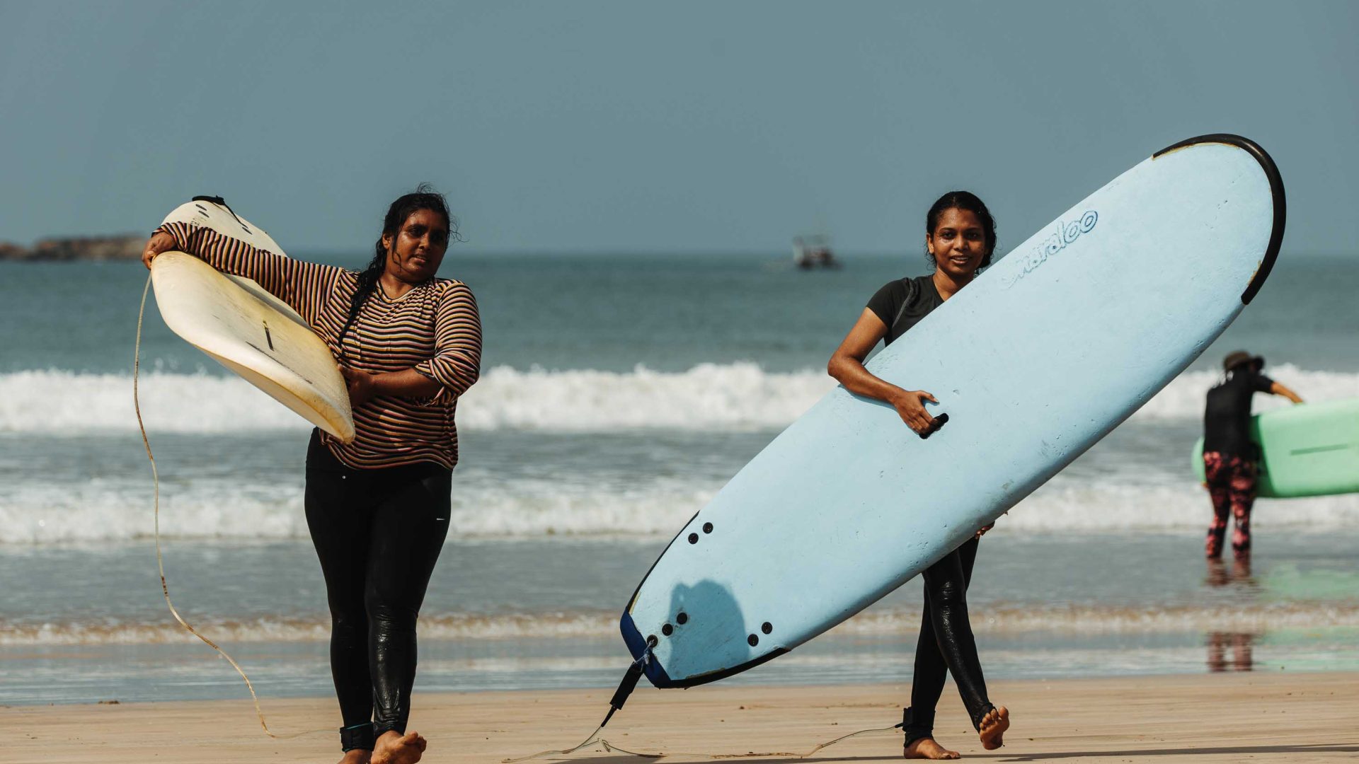 Women smile as they walk along the beach with their surf boards.