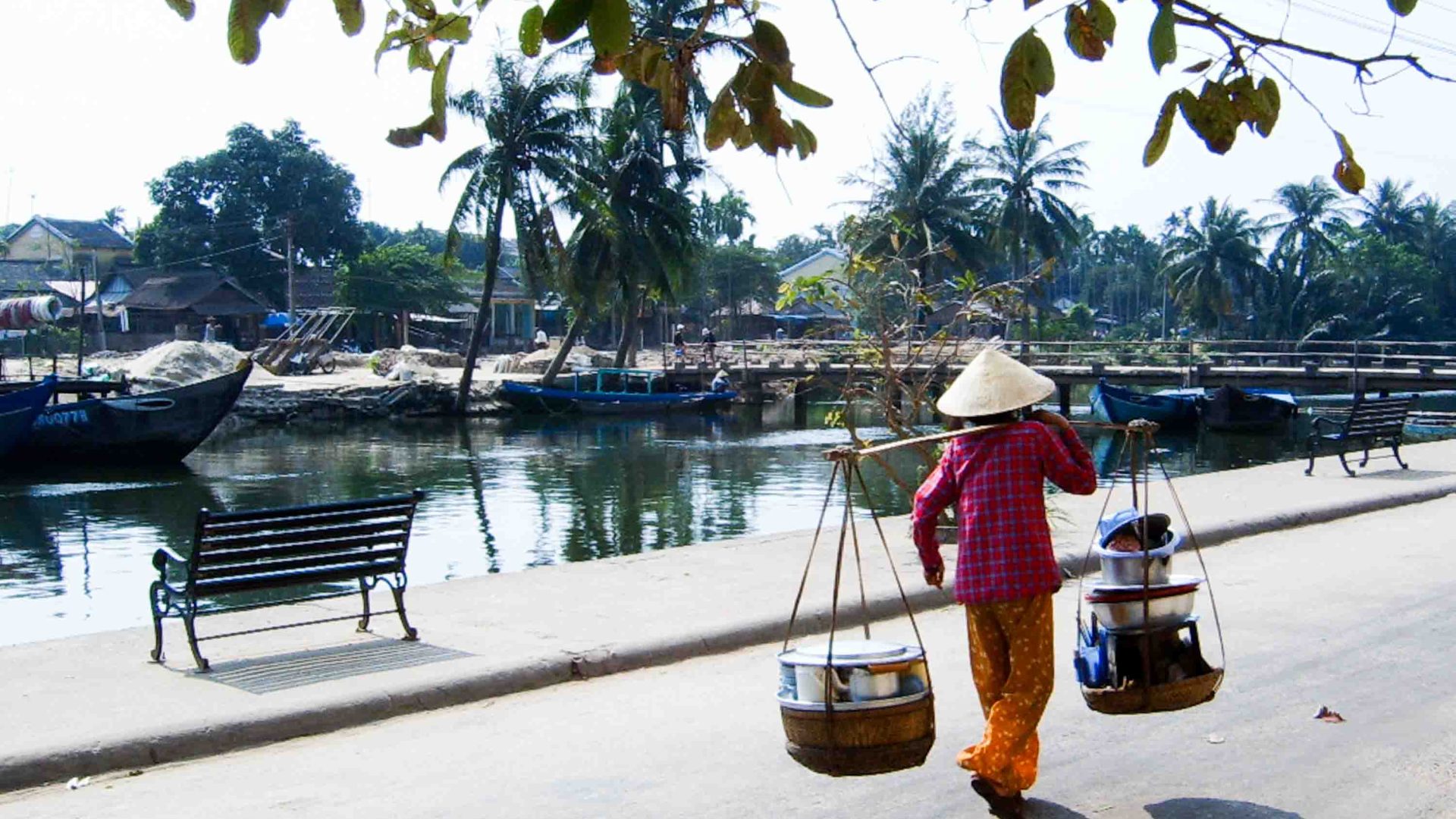 A woman carries two baskets. She wears a conical hat.