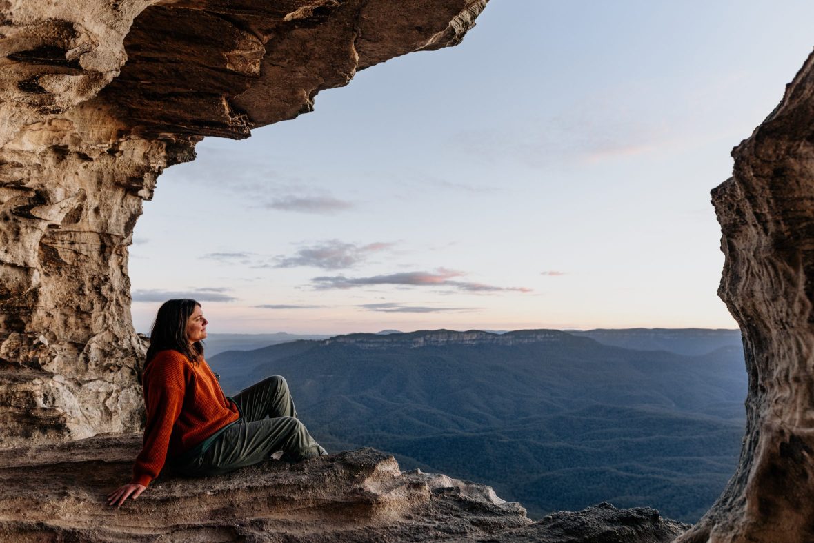 woman sits at cliff edge looking over mountain view