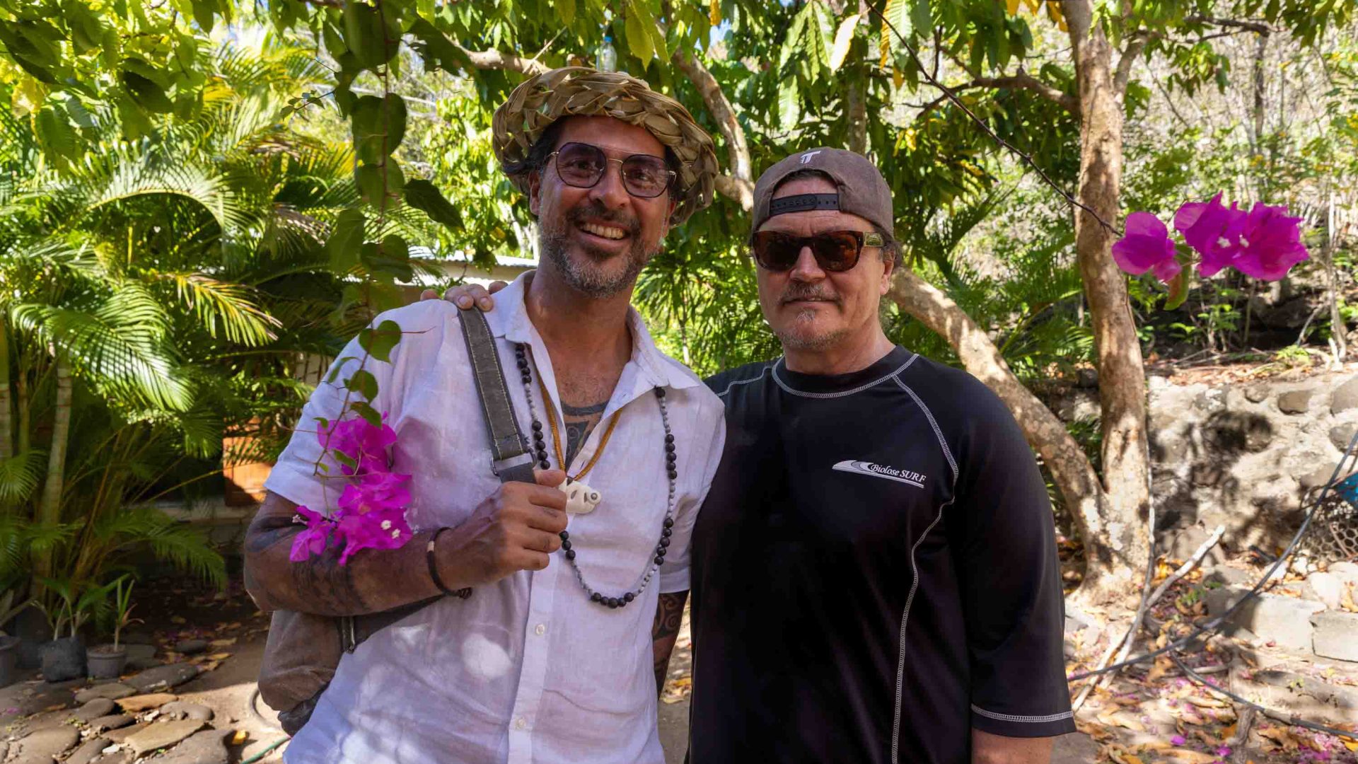 Two men smile under some trees. They hold purple flowers.