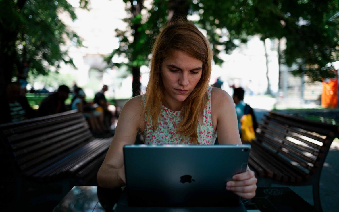 A woman looks at an iPad while seated in a park. There are other people in the background.