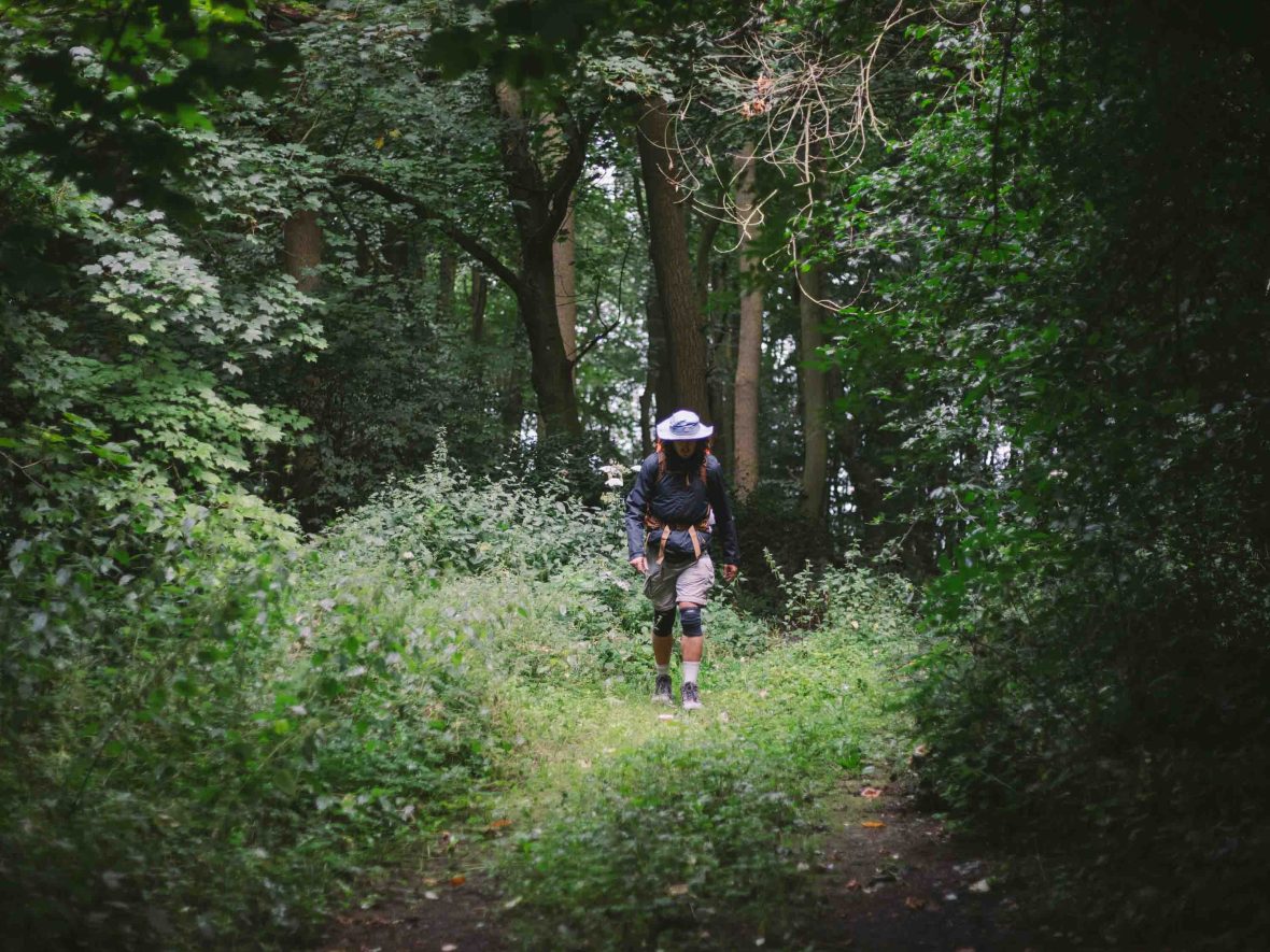 A man in a hat walks along a narrow path through trees.