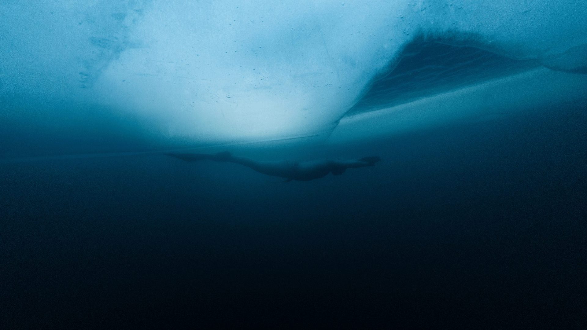 Side view of a freediver swimming under a thick sheet of ice. An escape hole shaped like a triangle is to the right.